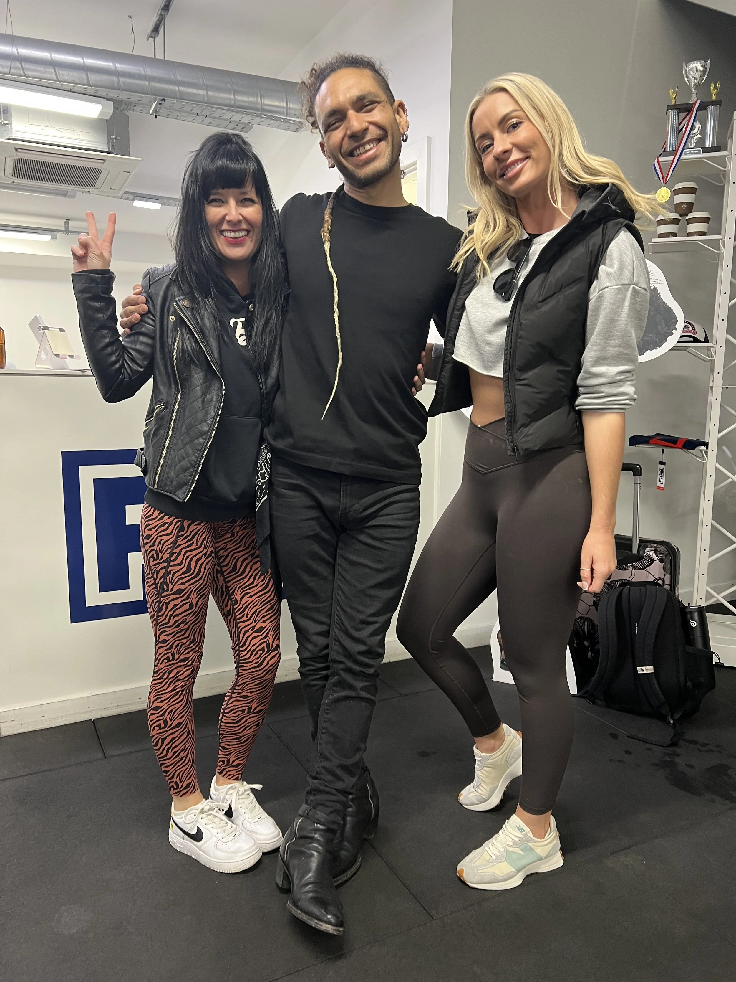 Three people standing together indoors, smiling and posing for a photo. One woman with black hair and a leather jacket, another woman with blonde hair and athletic wear, and a man with short dreadlocks wearing a black T-shirt. The background shows tr