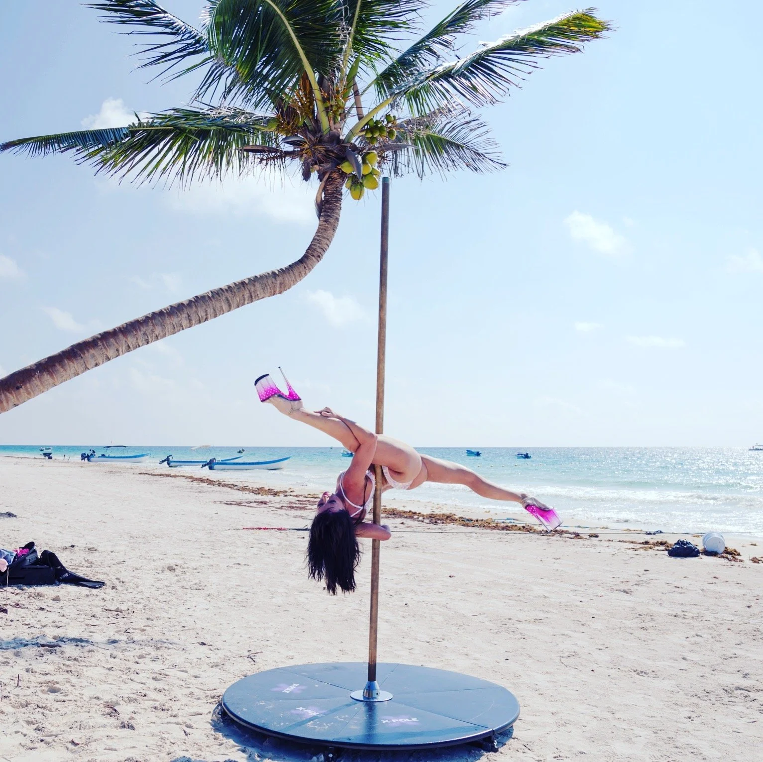 Person performing pole dance on a beach with palm tree and ocean in background, wearing pink shoes.
