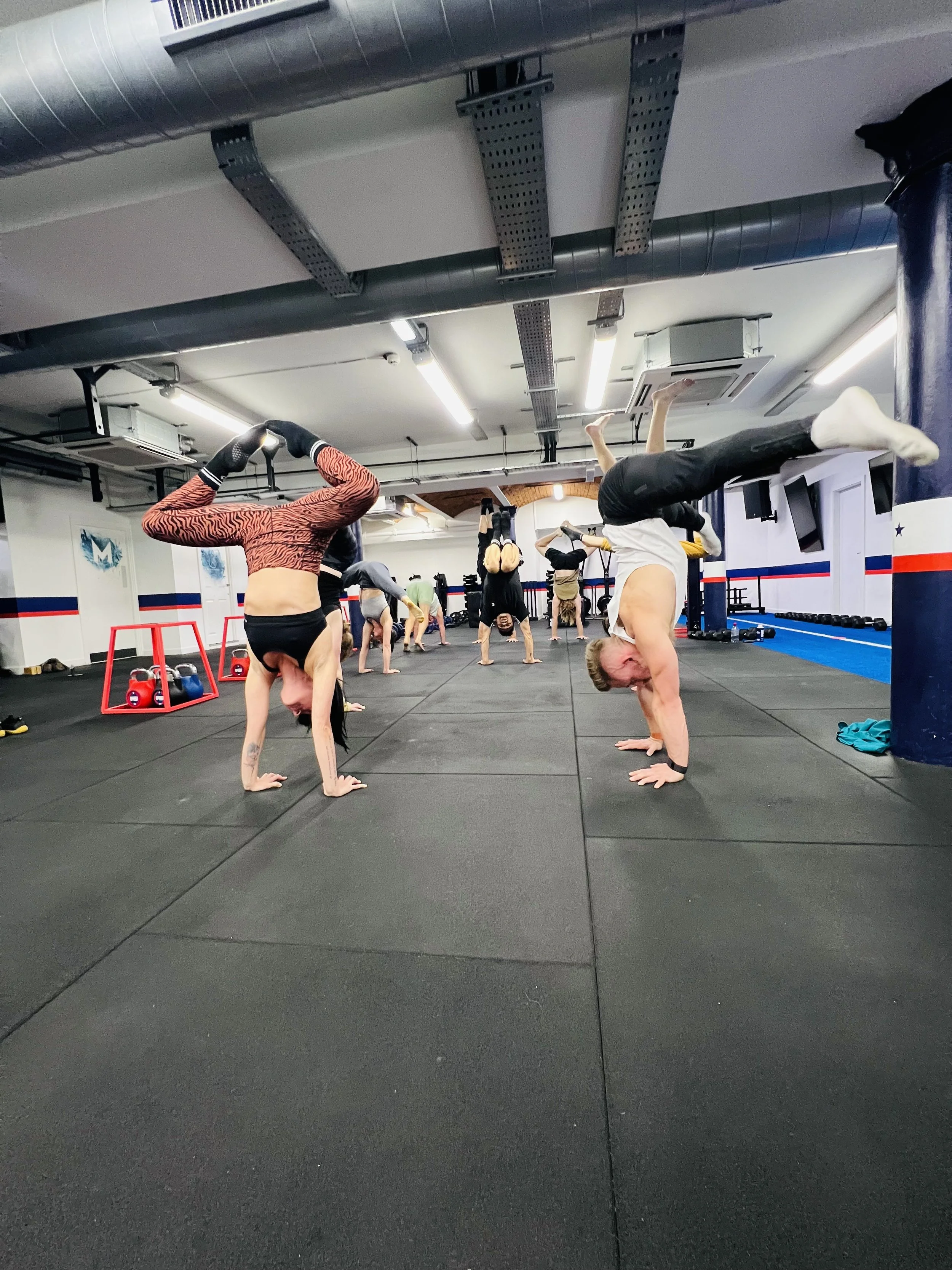 People practicing gymnastics or acrobatics in a gym, performing handstands and various balance poses on a black mat floor. The room has gym equipment and overhead lighting.
