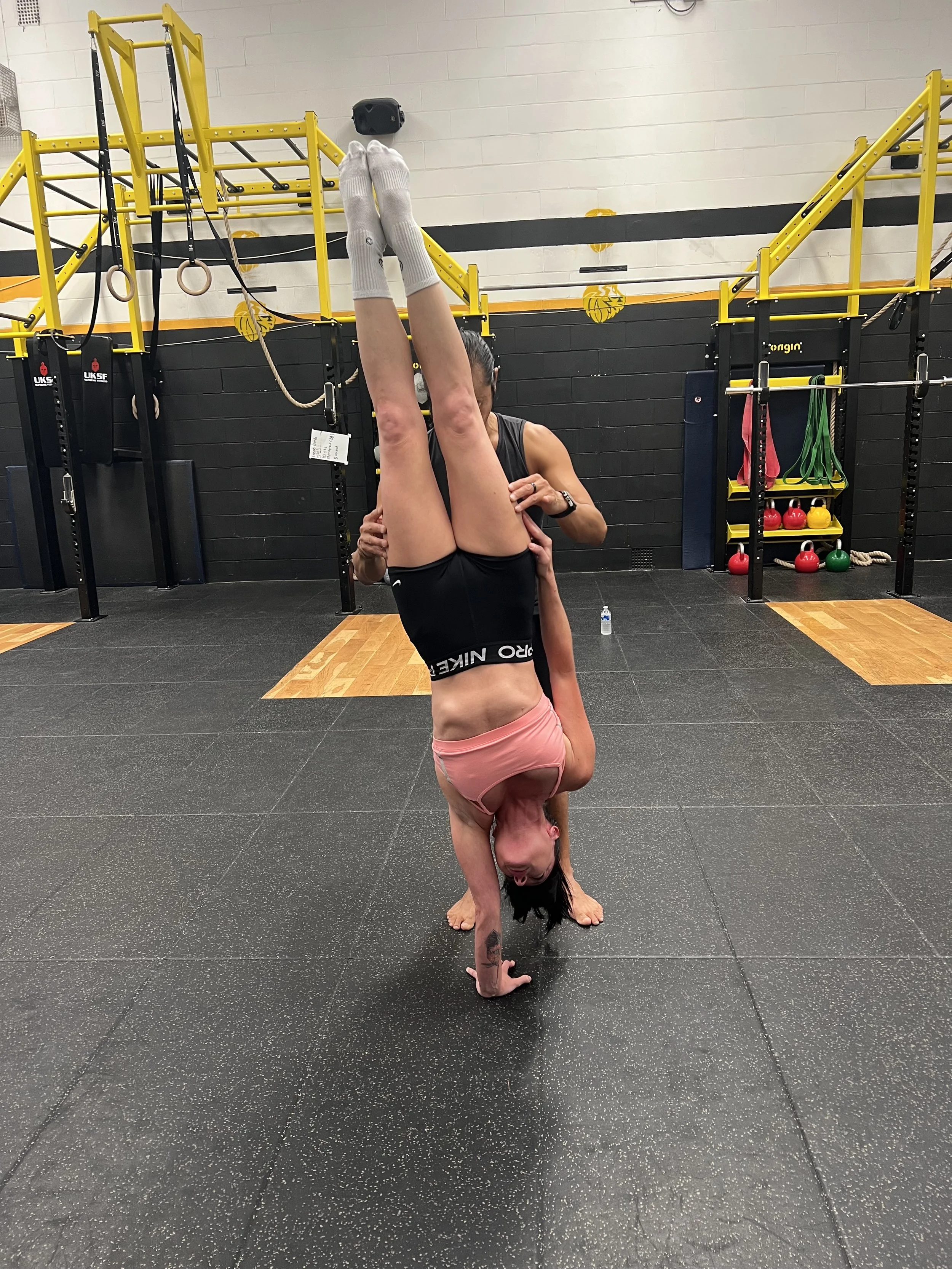 Two women practicing acro yoga in a gym, with one woman balancing upside down on her hands and shoulders while the other supports her legs.