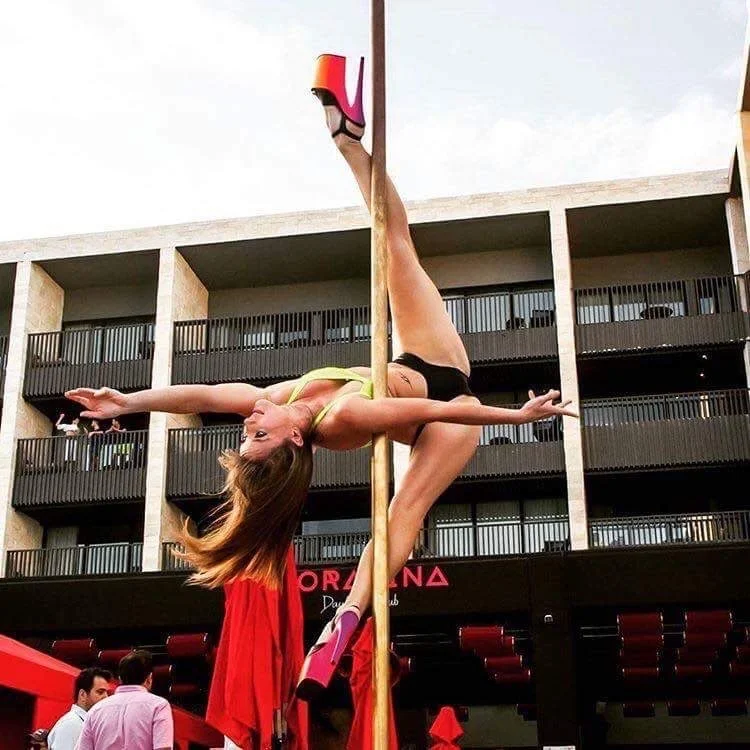Person performing a pole dance trick outdoors at a venue, wearing high heels and athletic wear, with a building in the background.