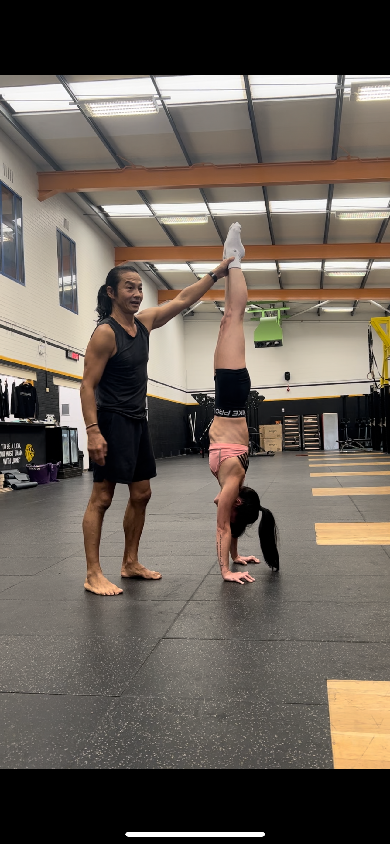A woman performing a handstand with assistance in a gym, guided by a man holding her feet upright.