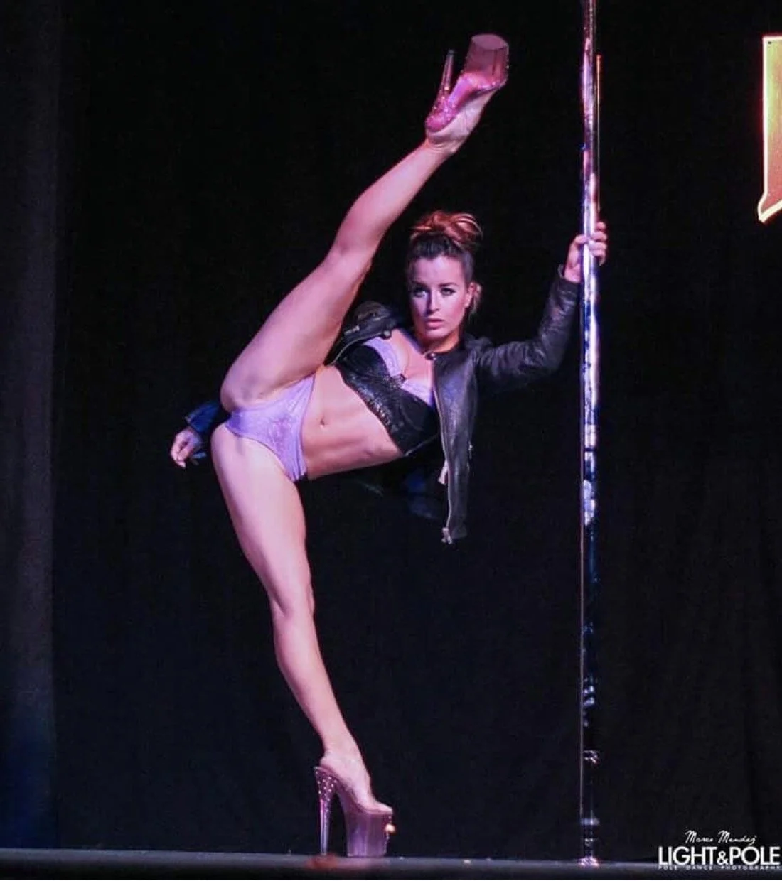 Pole dancer performing with one leg extended, wearing high heels and a jacket.
