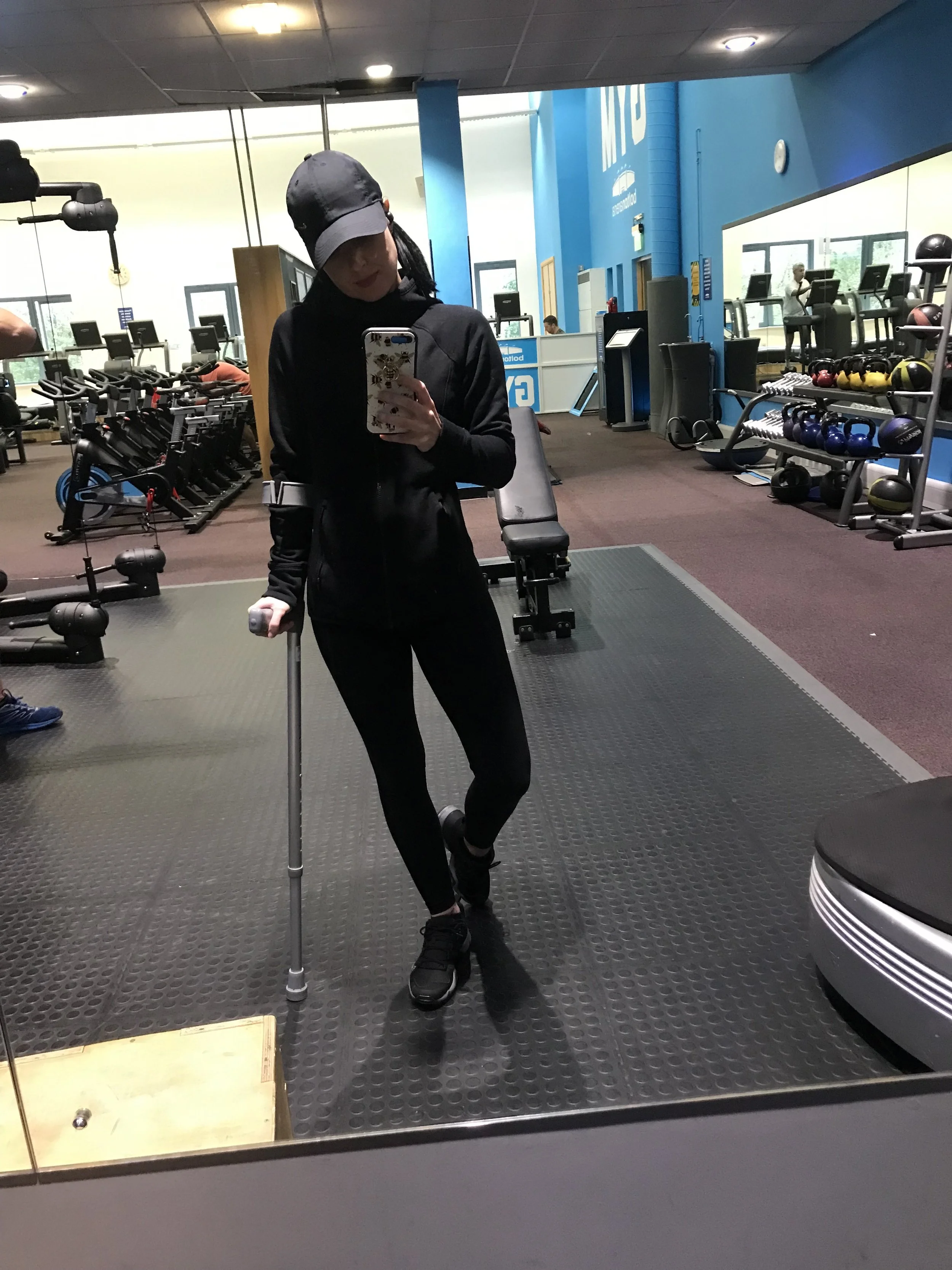 A woman taking a mirror selfie in a gym, wearing black workout clothes and a cap, holding a small white object in one hand and a phone in the other.