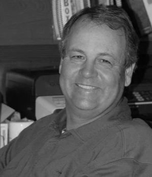 A smiling man with short hair, wearing a collared shirt, sitting in an indoor setting.
