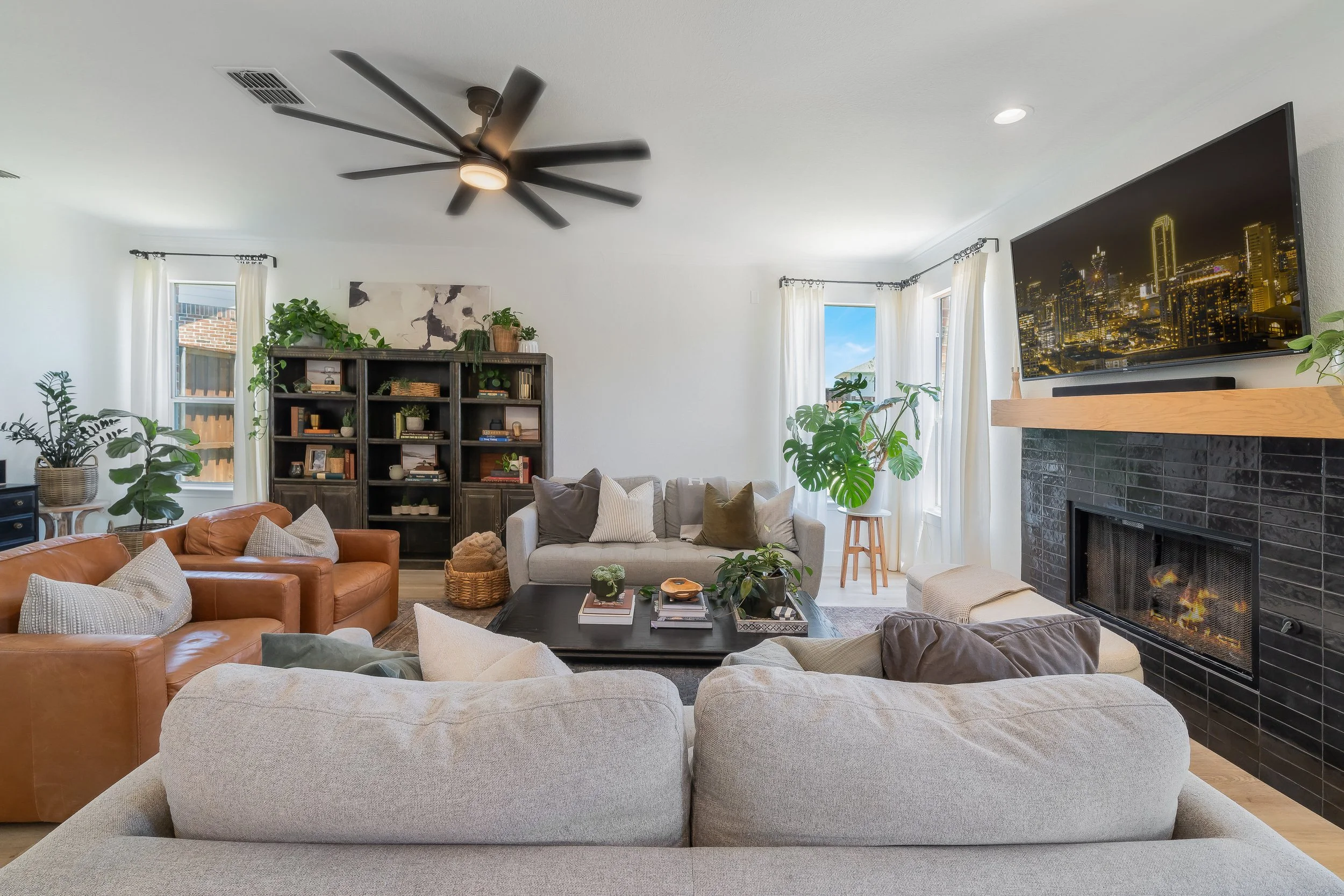 Living room with beige, gray, and brown sofas, a black coffee table, a dark bookshelf, a black brick fireplace with a mounted TV, and large windows with white curtains, decorated with indoor plants and cityscape at night on the TV screen.