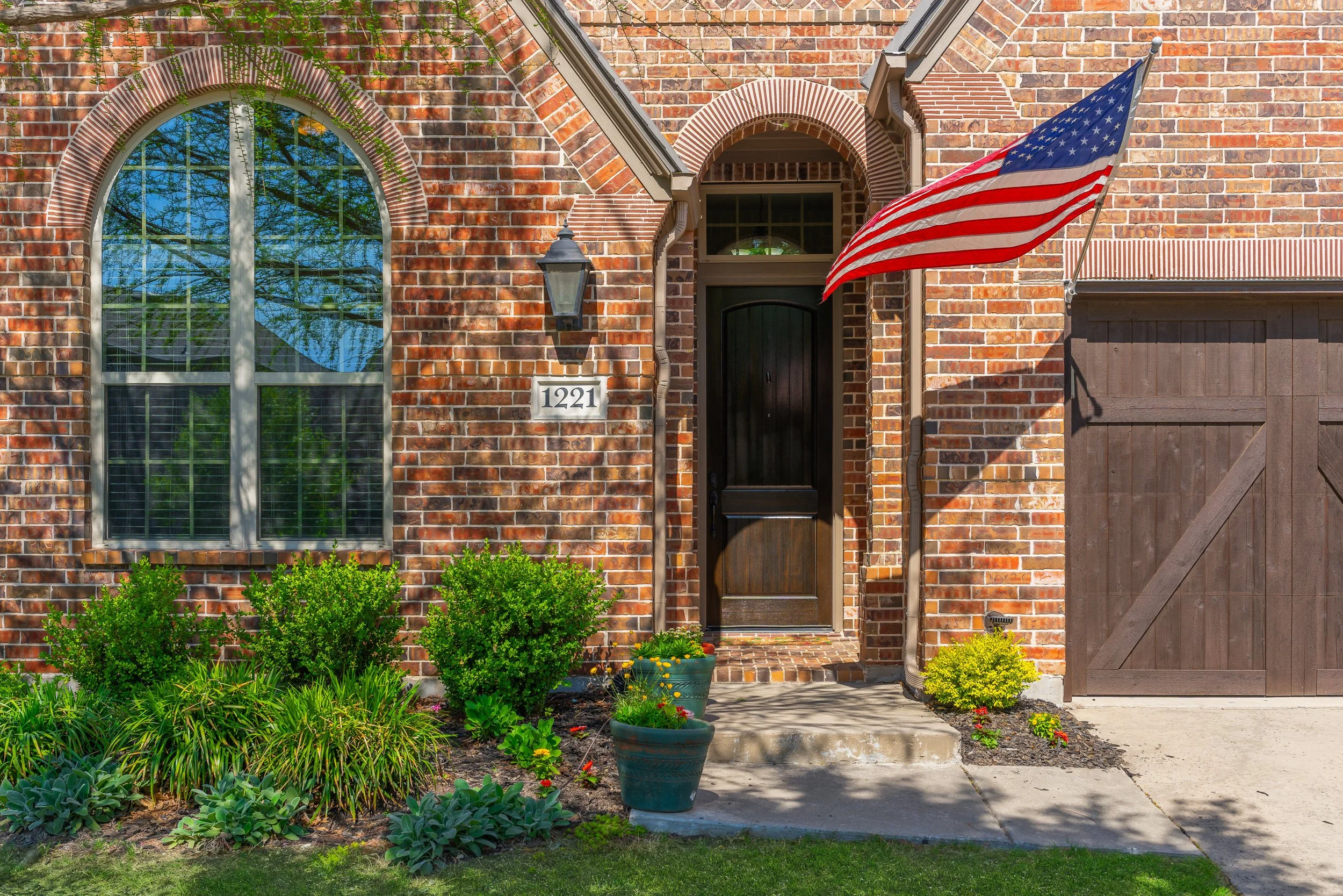 Brick house entrance with a black door, a large arched window, outdoor light fixture, US flag, house number 1221, and a garden with green shrubs and potted plants.