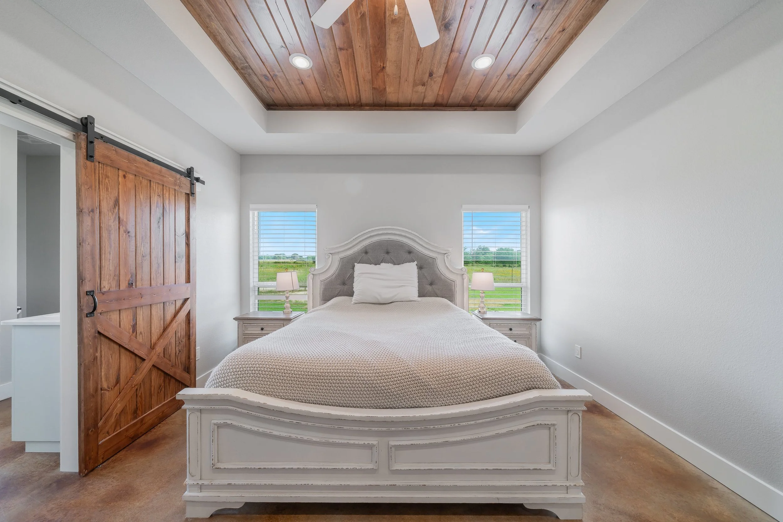 Bedroom with white wooden bed, beige bedding, two nightstands with lamps, sliding barn door, and windows showing green landscape outside.