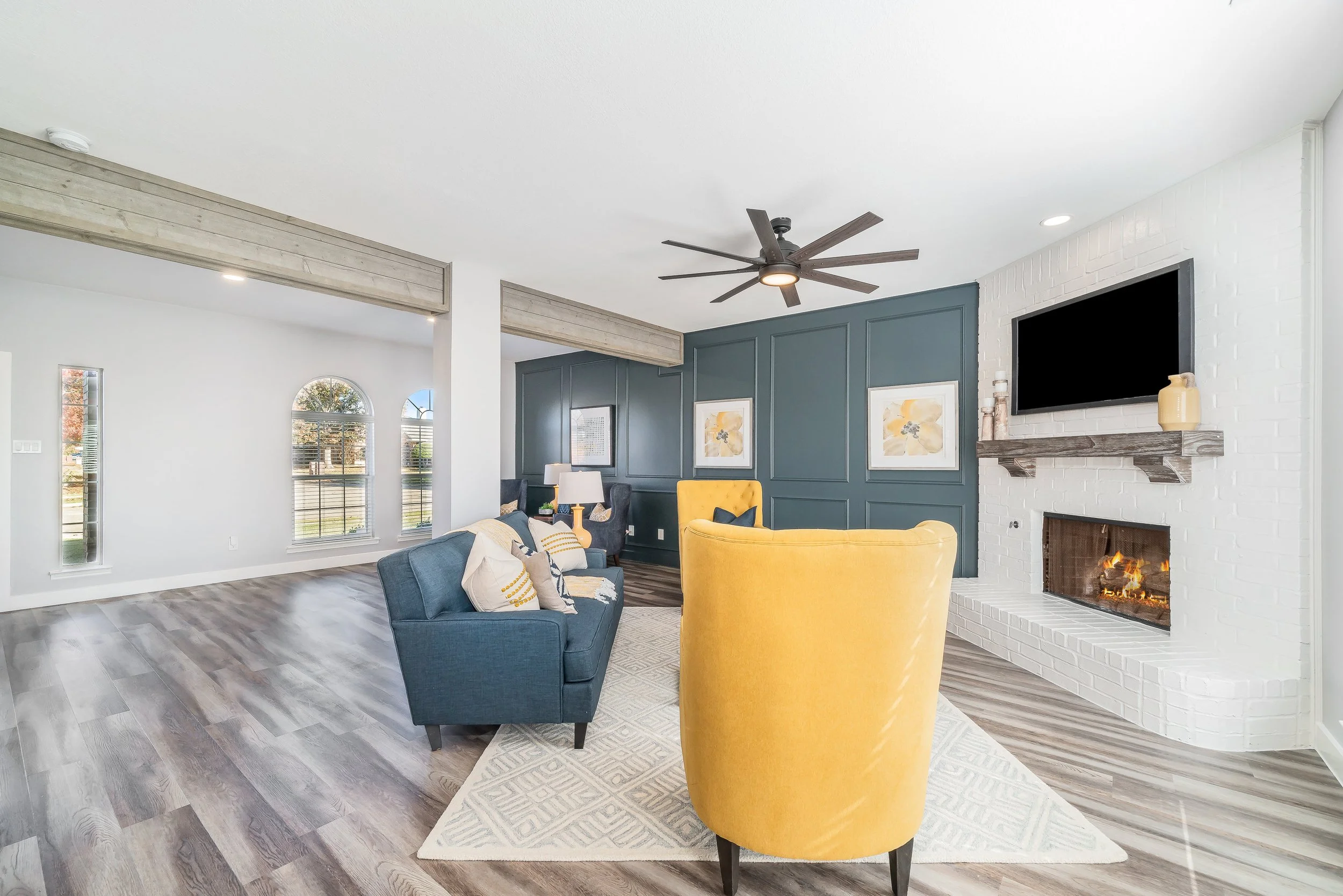 Living room with a yellow armchair, a dark blue sofa, a gray armchair, a fireplace with a television above, a ceiling fan, and artwork on the blue accent wall.
