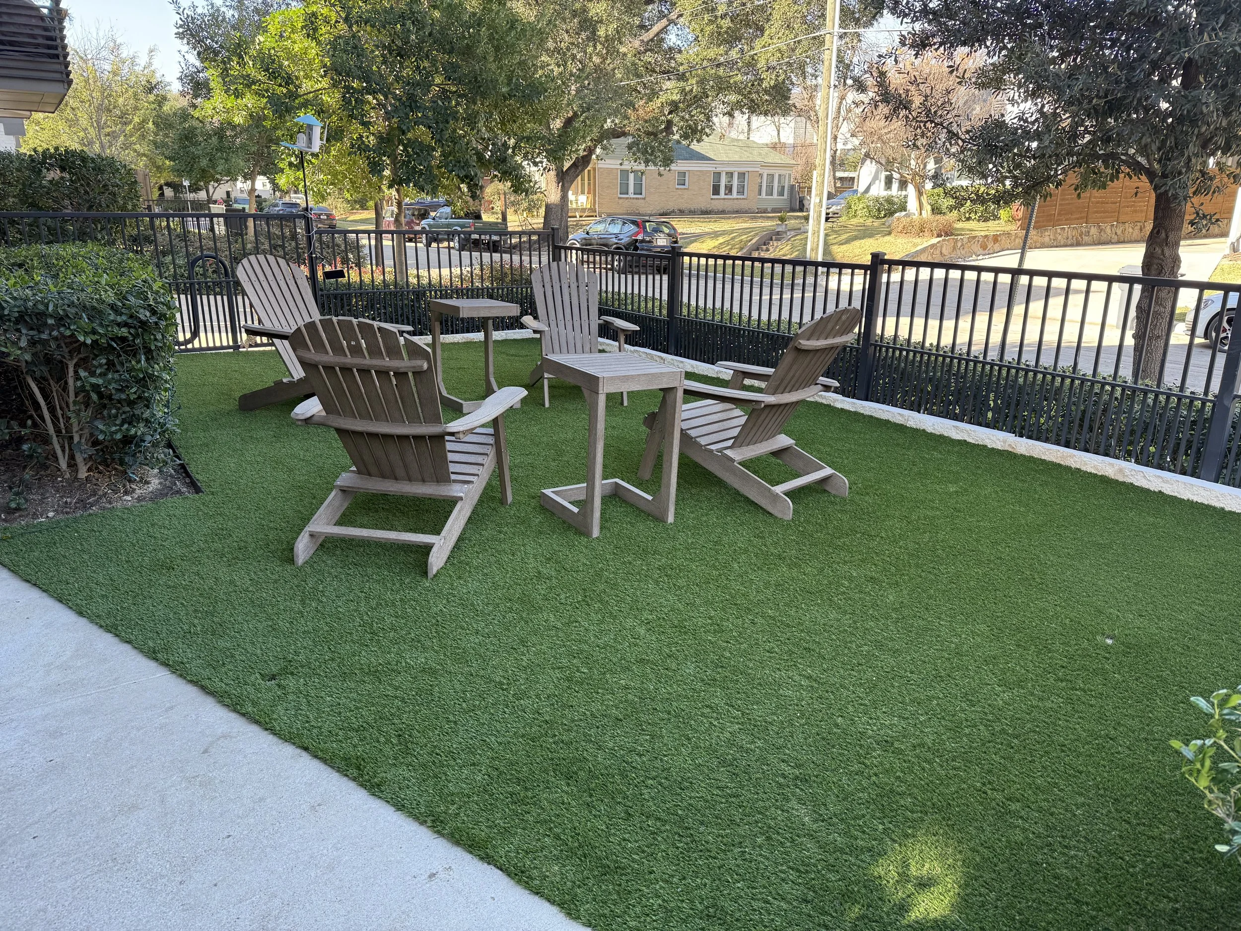 Outdoor patio area with four wooden Adirondack chairs and two small tables on a patch of artificial grass, enclosed by a black metal fence, with trees and residential houses in the background.