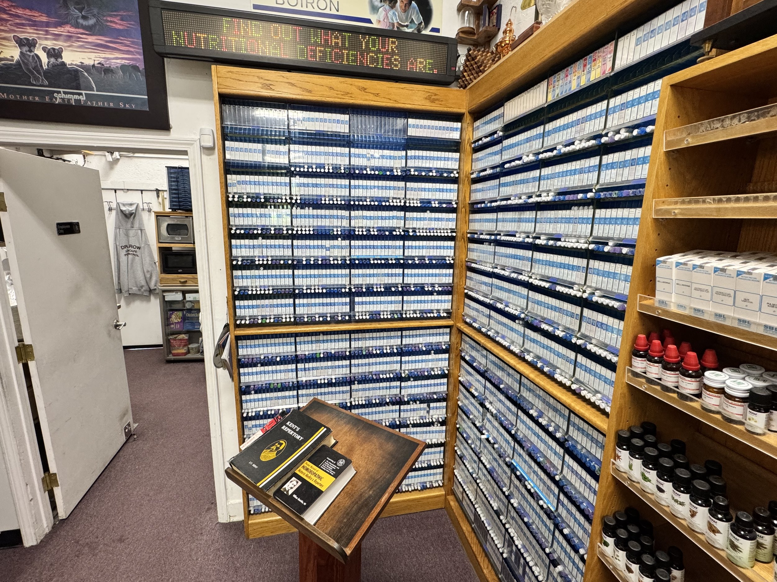 An interior of a pharmacy or medical supply store with shelves filled with medication blister packs. There is a small table in front with a couple of notebooks and a book. A digital signboard above reads, 'Find out what your nutritional deficiencies are.'