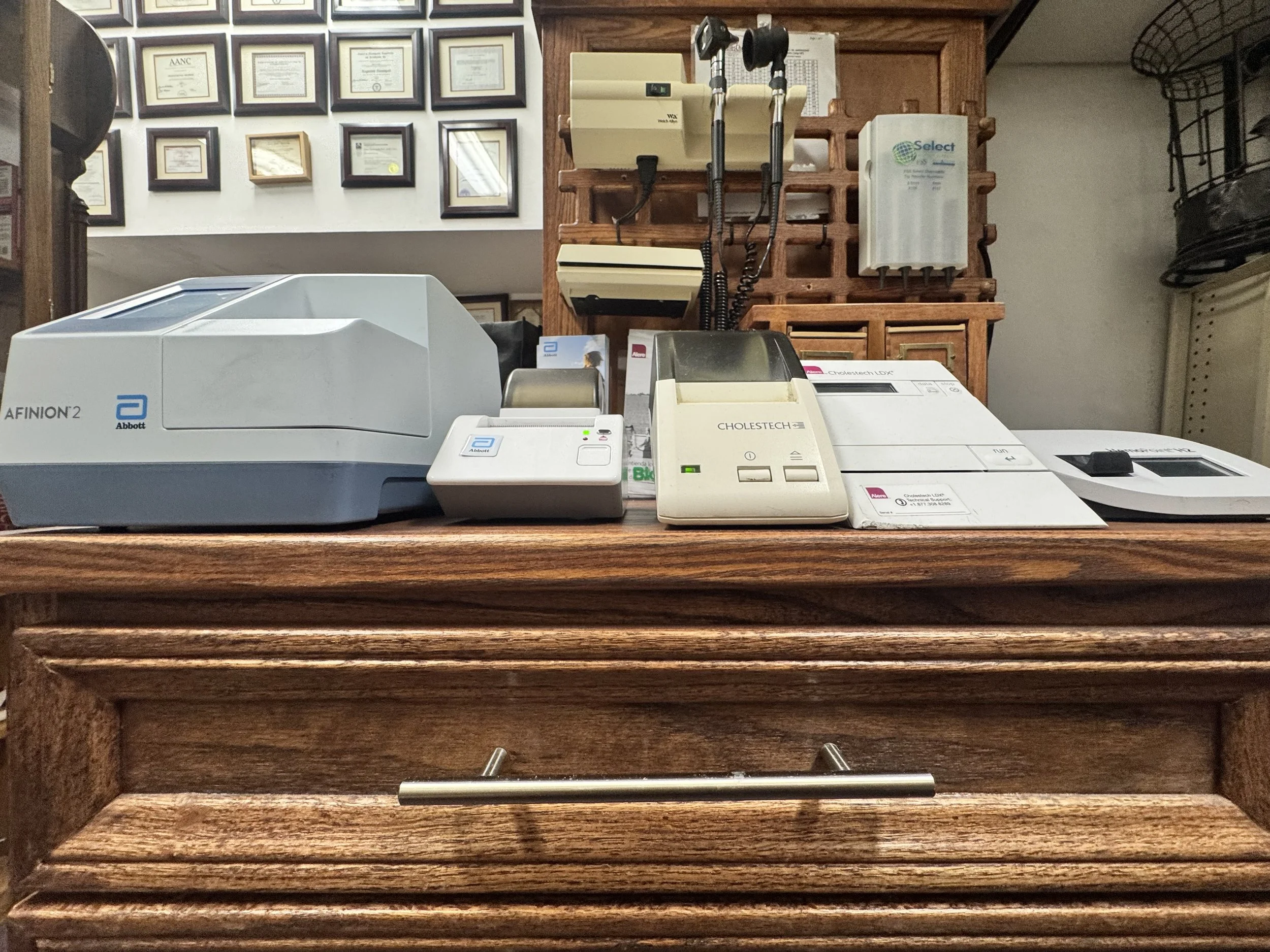 Medical equipment and devices on a wooden countertop in a clinic, including an Abbott Afinion 2 analyzer, a small printer, a Cholestech testing device, and other diagnostic tools.