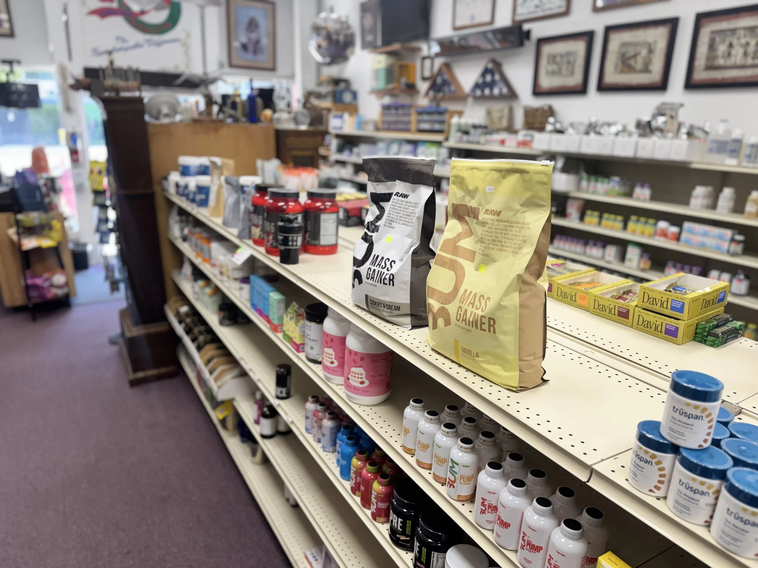 Store aisle with dietary supplements and protein powders on shelves, including bags of RAW mass gainer and containers of powder or capsules.
