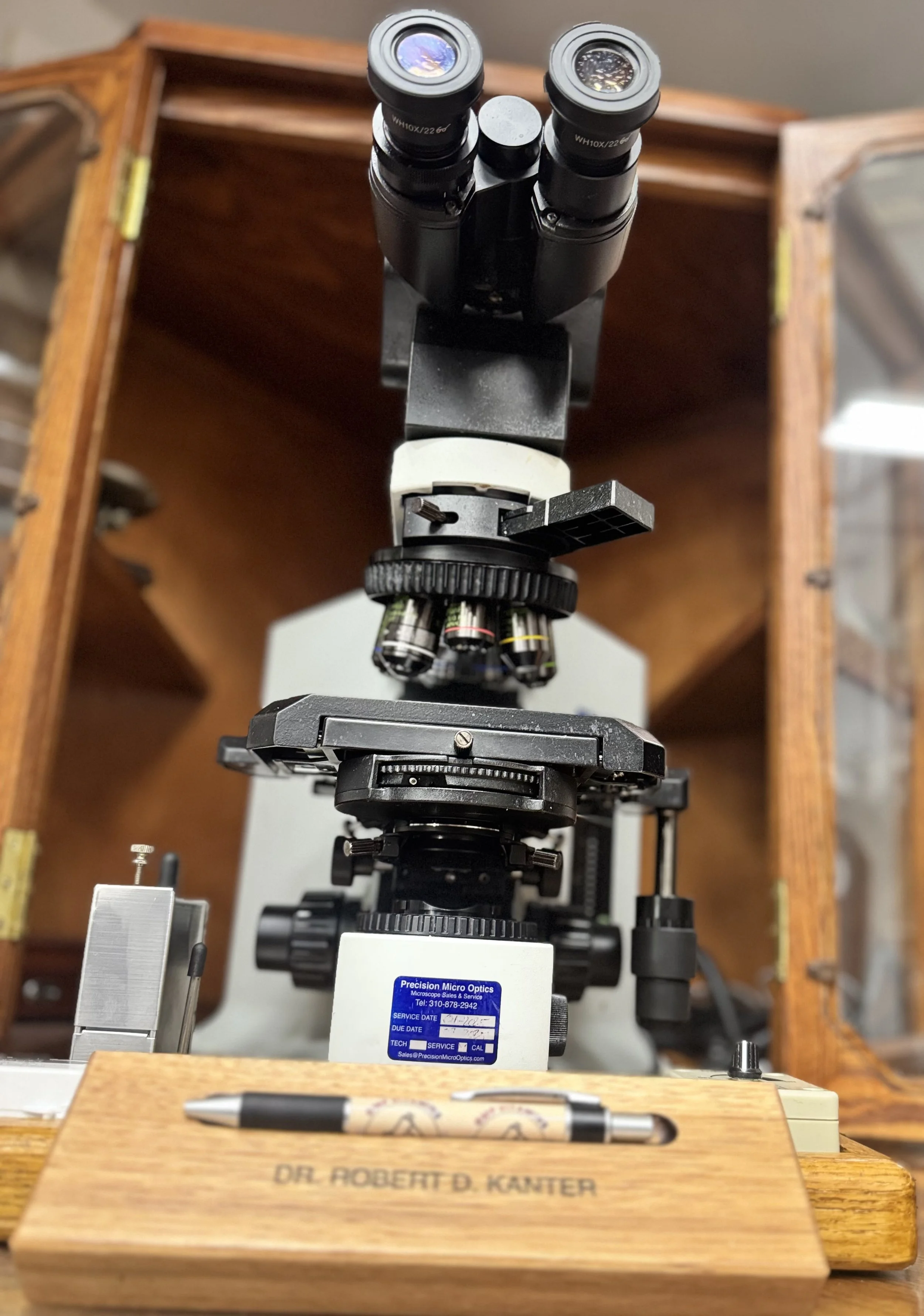 Microscope on a wooden table with a pen in the foreground, engraved with the name 'DR. ROBERT D. KANTER', in a lab setting with wooden cabinets in the background.