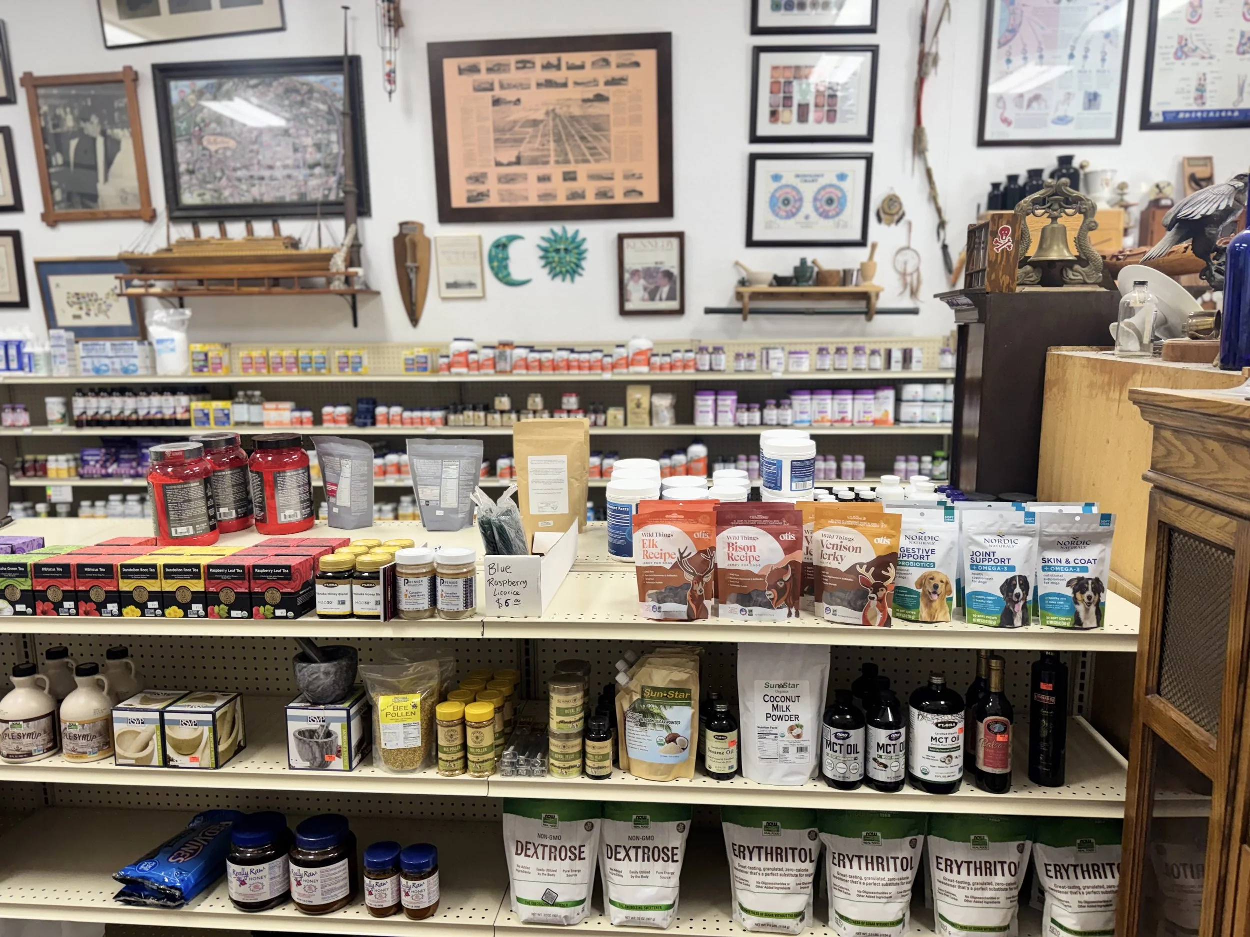 Interior of a pet supply store with shelves stocked with various pet supplements, food, and health products. The back wall displays framed pictures and decorative items.