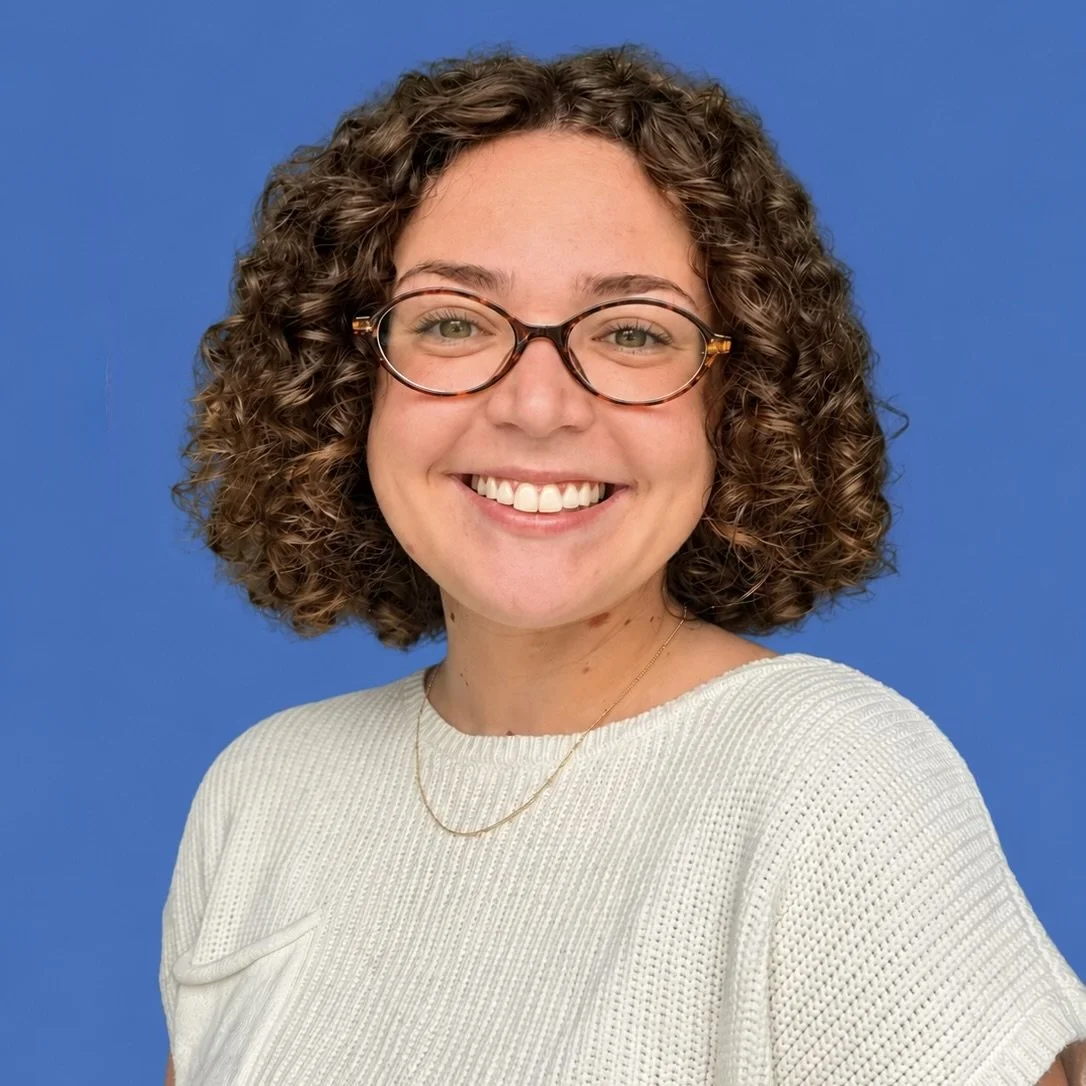 A smiling woman with curly hair and glasses sitting outdoors during the daytime.