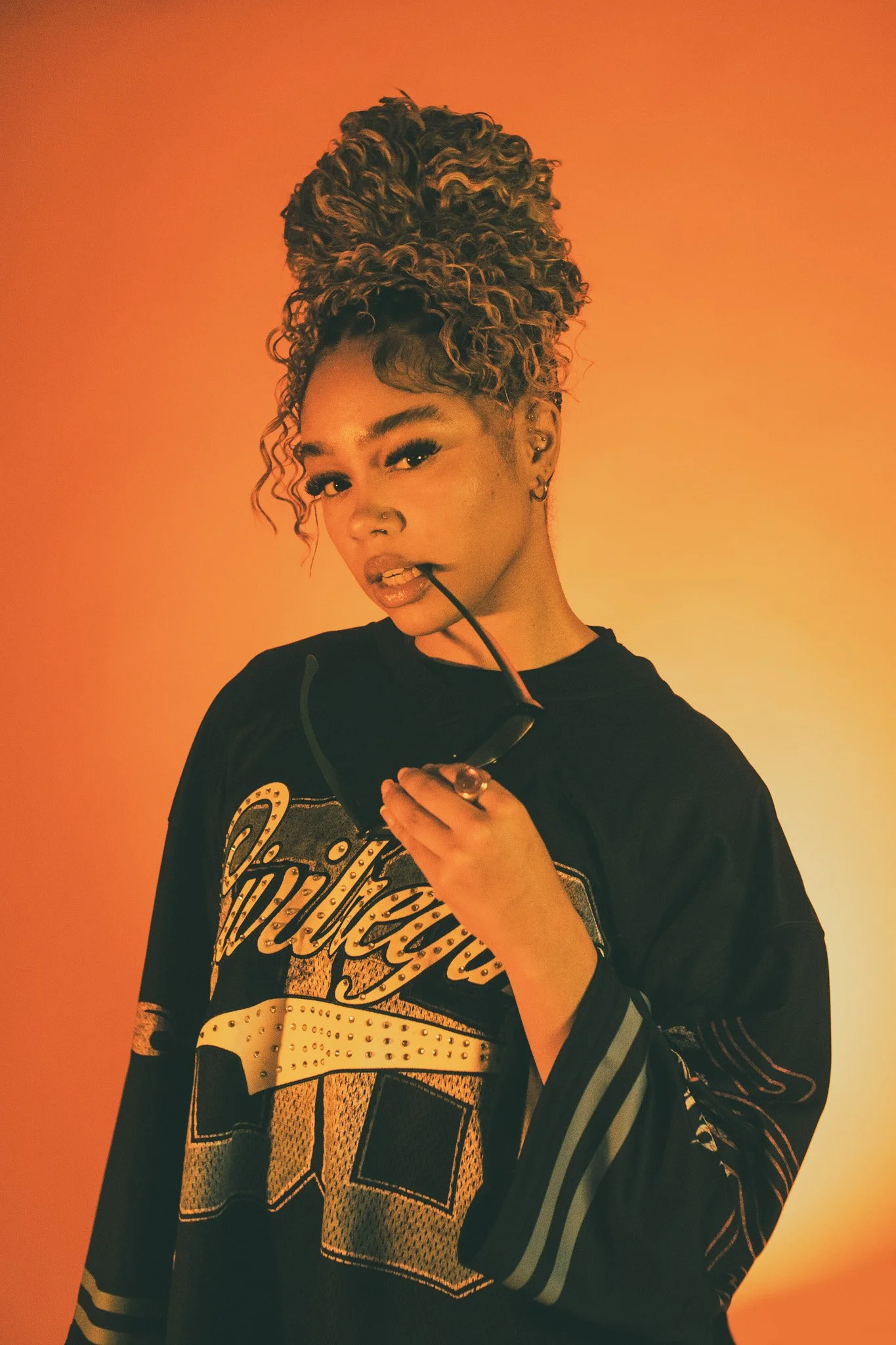 Young woman with curly hair in a topknot, holding sunglasses, against warm orange background.