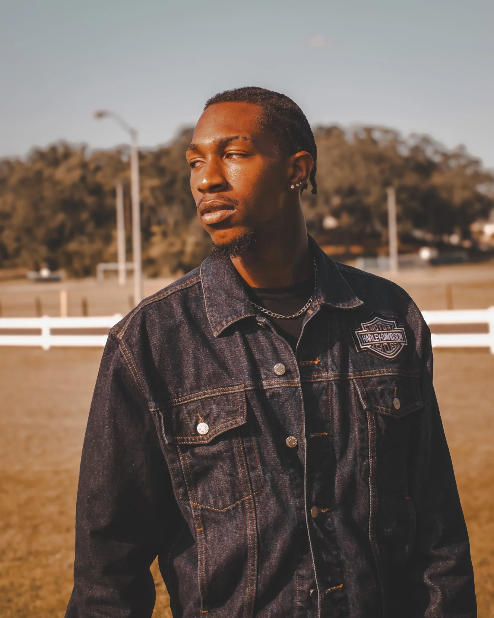 A young man with braided hair and earrings wearing a black Harley Davidson denim jacket standing outdoors during daytime.