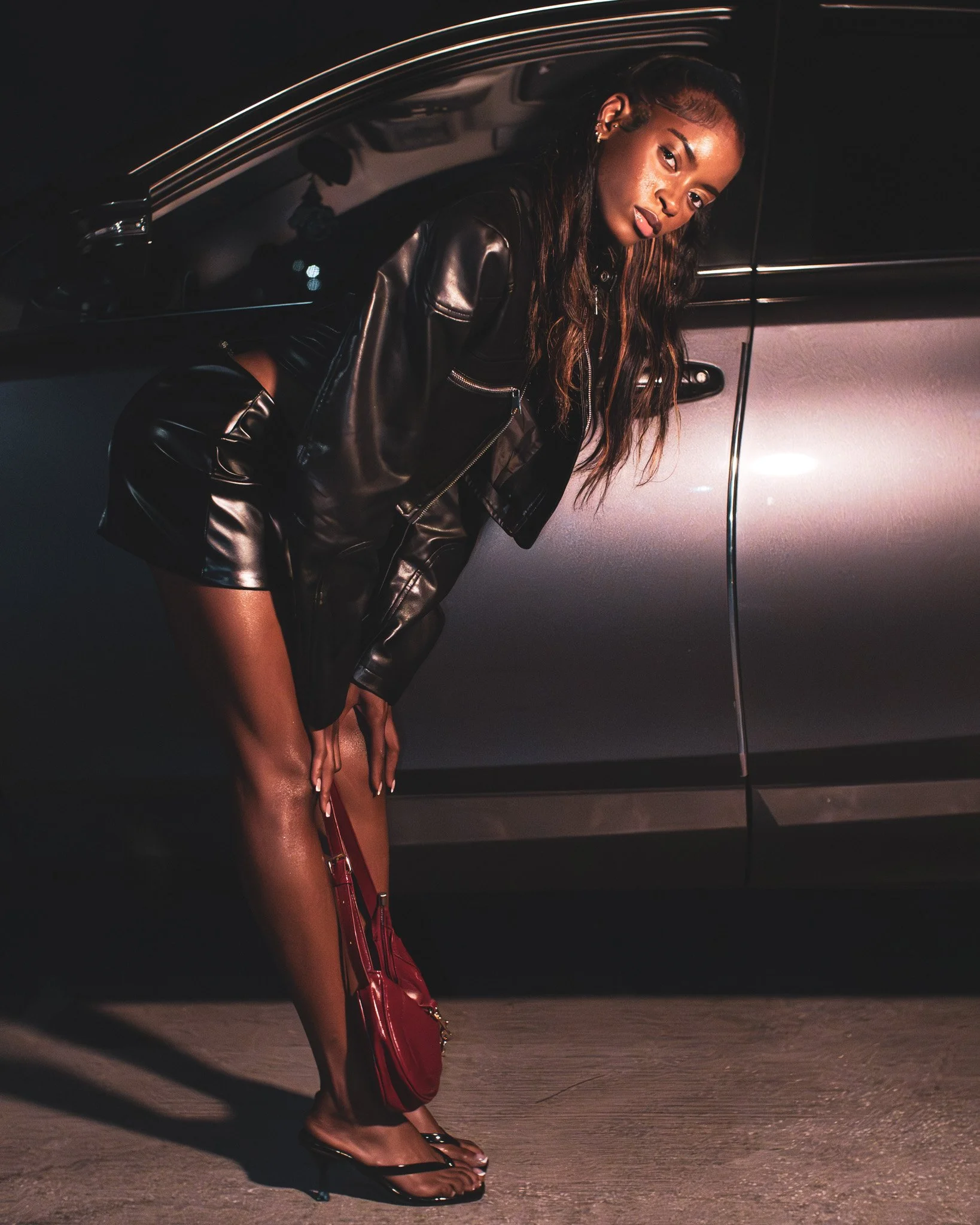 A woman leaning against a car at night, wearing a black leather jacket, matching shorts, and black heeled sandals, holding a red purse.