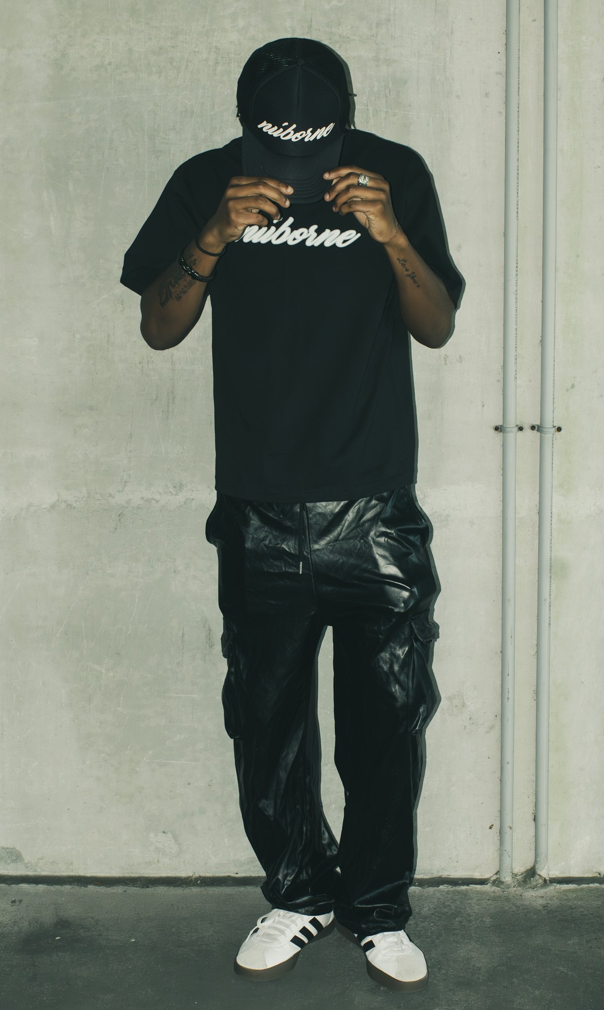 Person wearing a black hat and black t-shirt with white cursive text, black shiny pants, striped sneakers, standing against a plain wall with visible pipes.