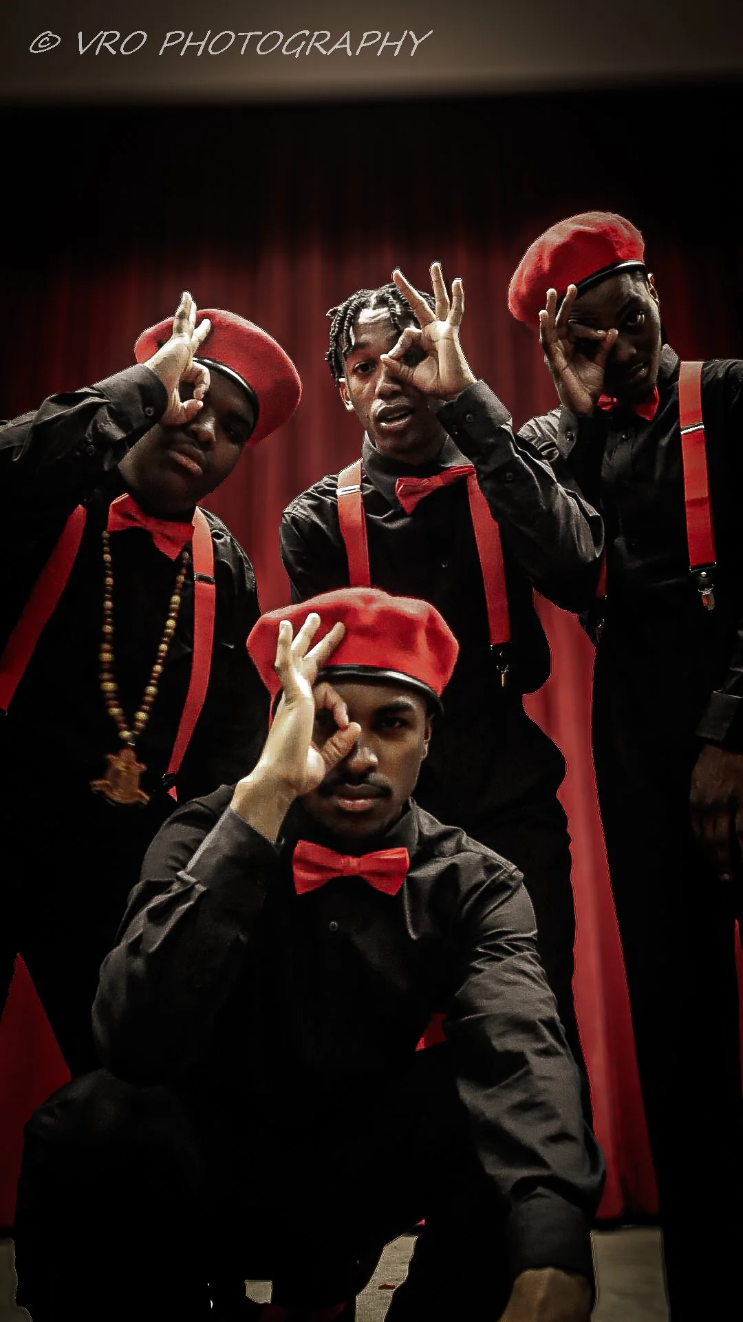 Four young men dressed in black shirts with red suspenders and red bow ties, wearing red berets, in front of a red curtain, making hand gestures to their eyes.