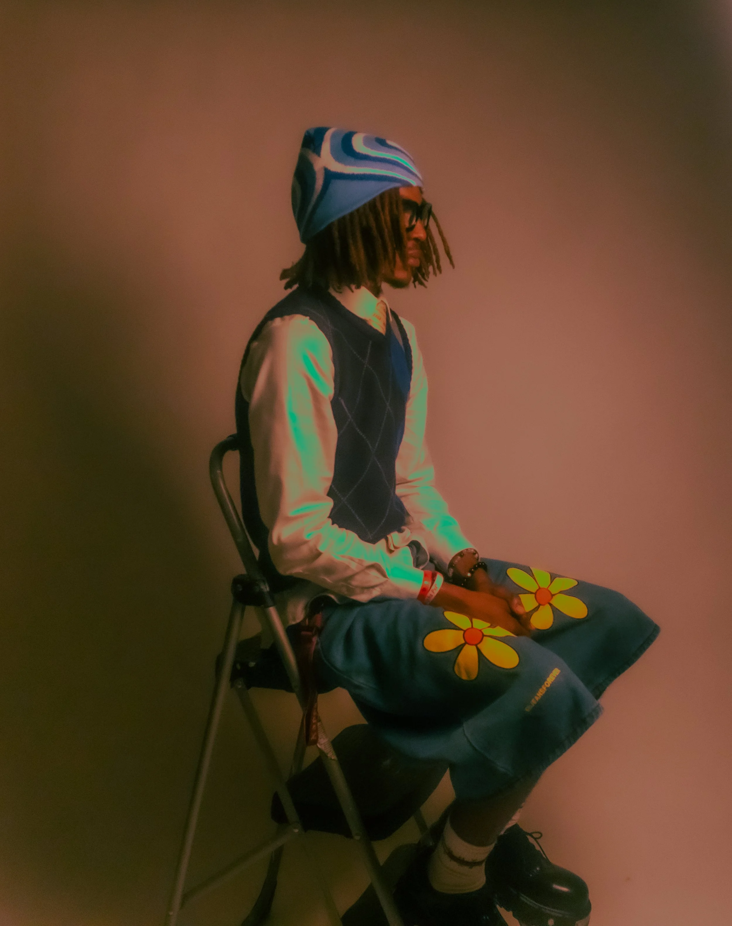 Person with dreadlocks wearing glasses, a blue patterned beanie, a navy blue vest over a light-colored shirt, and floral shorts, sitting on a folding chair against a plain wall.
