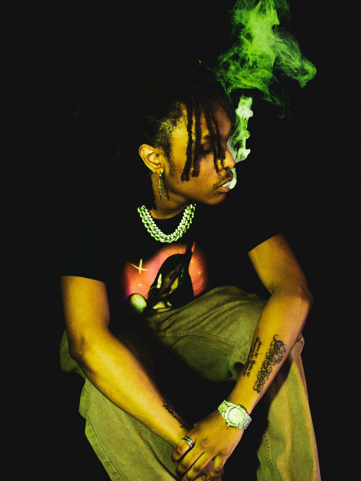 A person with dark skin and dreadlocks smoking, with green smoke around the face, wearing a pearl necklace, a wristwatch, rings, and tattoos on the arms, sitting against a black background.