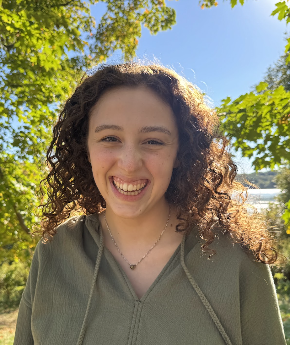 A young woman with curly hair smiling outdoors on a sunny day, surrounded by trees, wearing an olive green hoodie and a silver necklace.