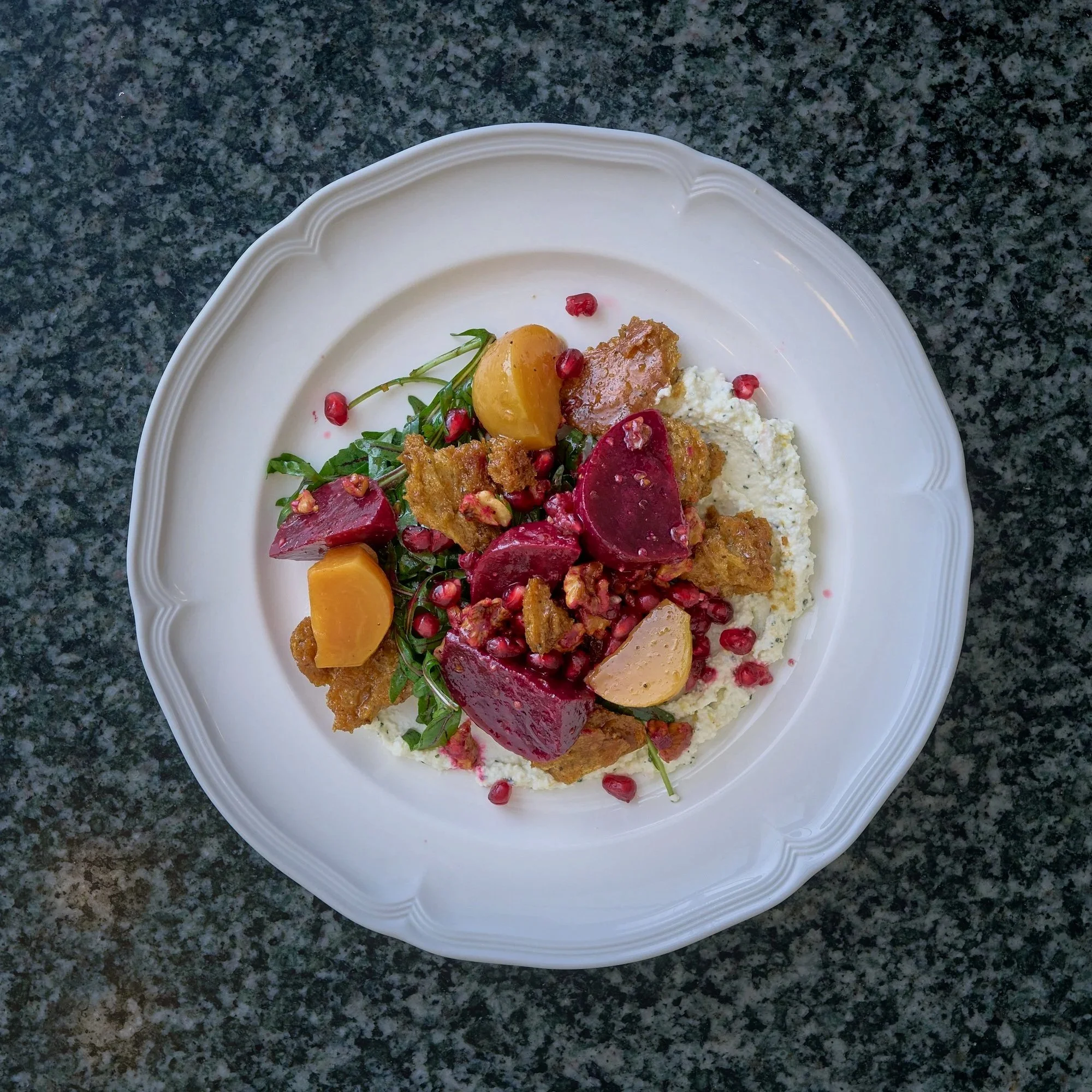 Close-up of a white porcelain plate with food, including sliced beets, potatoes, greens, and pomegranate seeds, on a dark granite countertop.