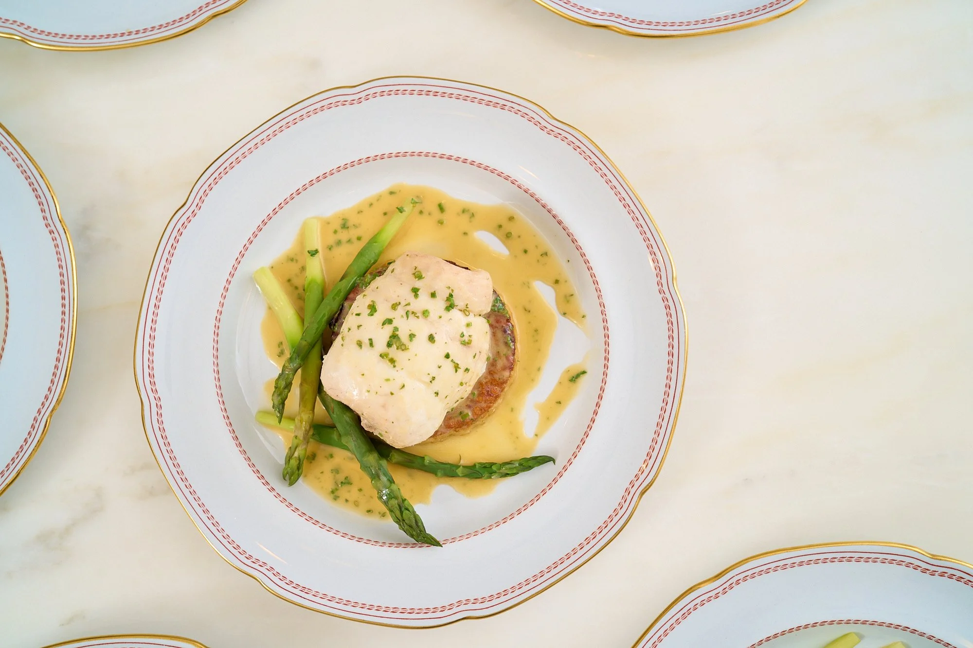 A plated dish with a piece of fish topped with herbs, served with green asparagus and a creamy yellow sauce, on a white plate with red and gold trim.