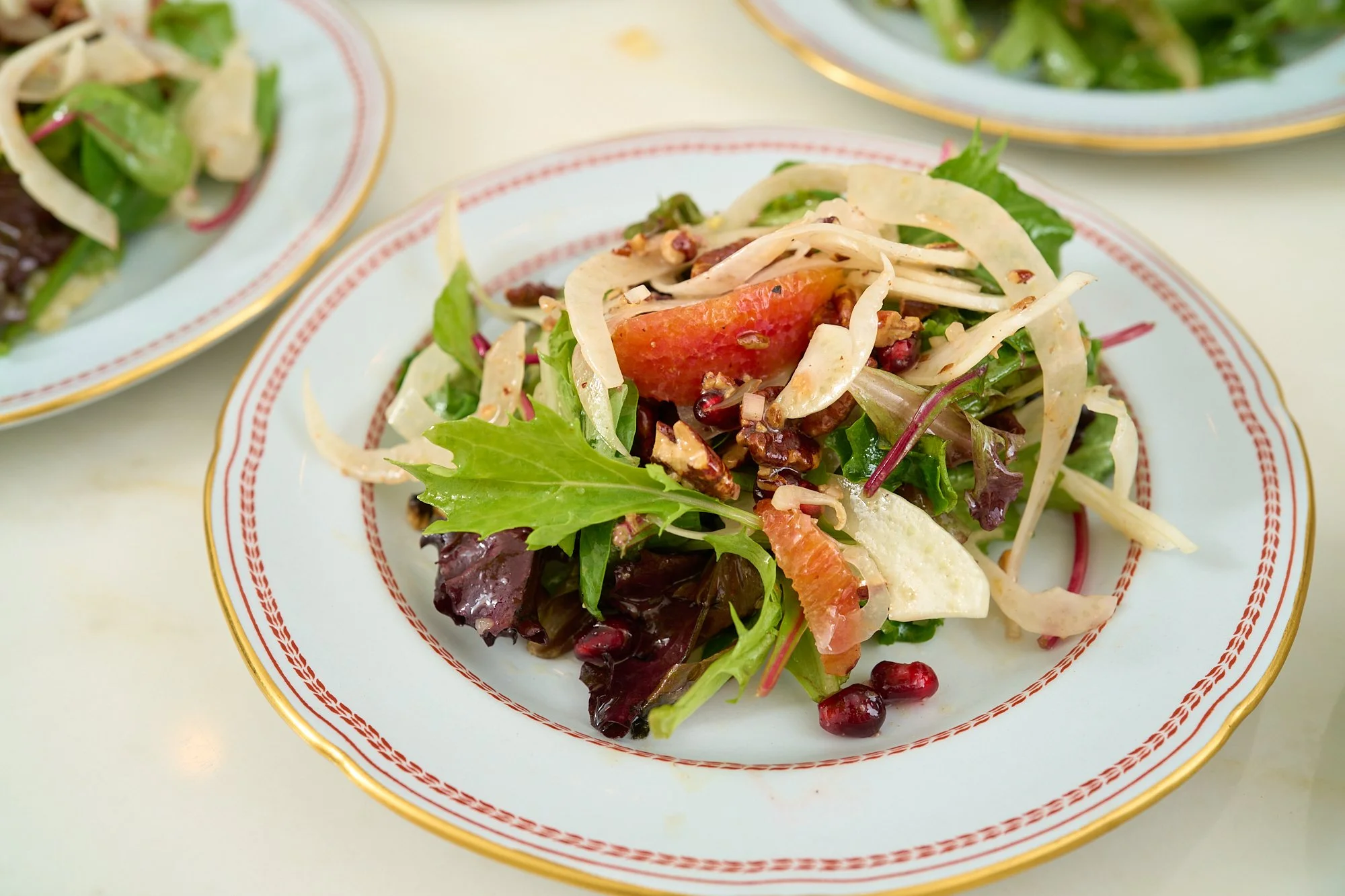 A plate of mixed salad with greens, sliced apple, pomegranate seeds, and nuts, served on a decorative white plate with a red and gold rim.