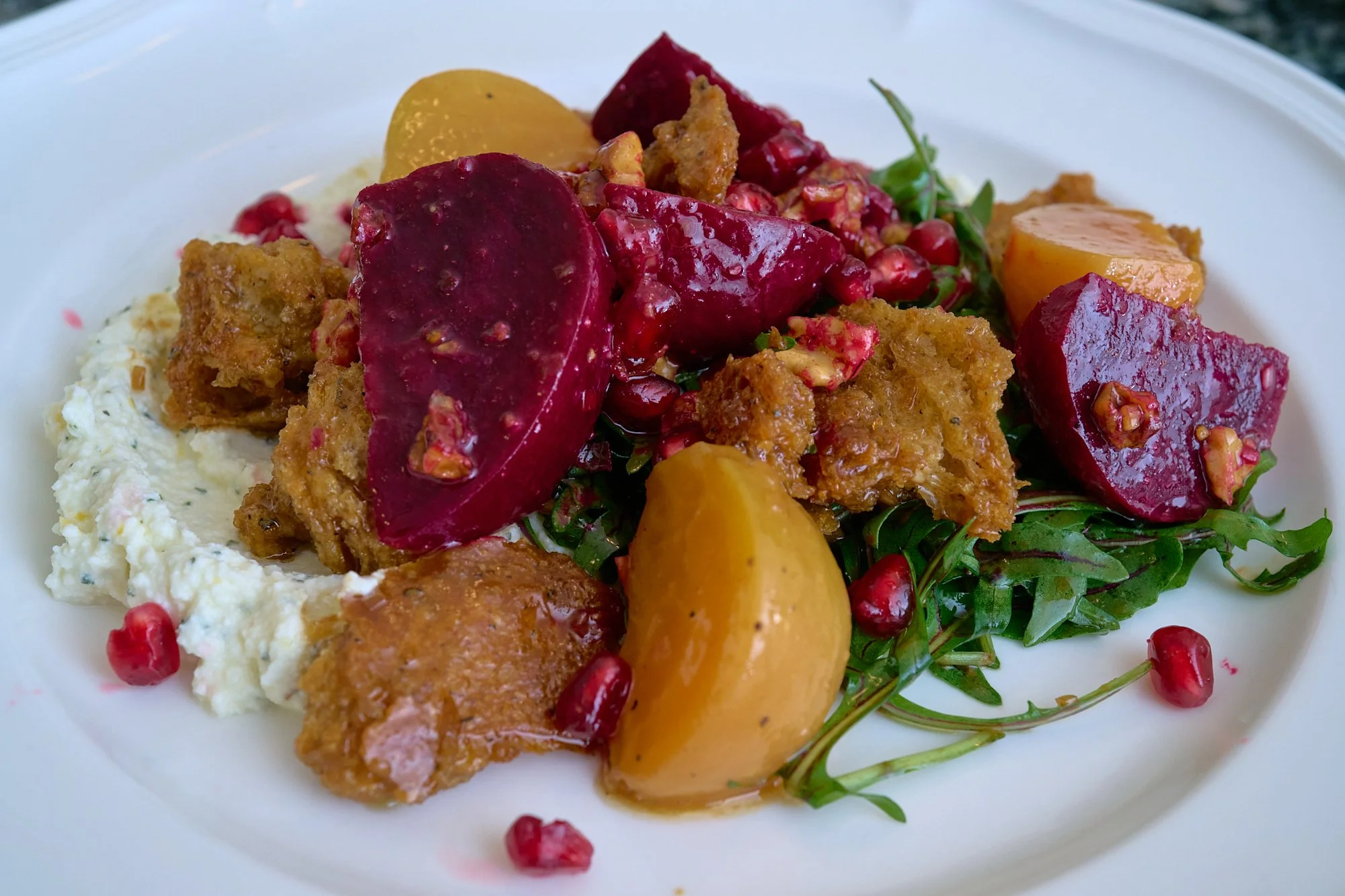 A plate of food with roasted beets, squash, chopped herbs, microgreens, roasted pieces of chicken, pomegranate seeds, and a bed of mashed potatoes with herbs.