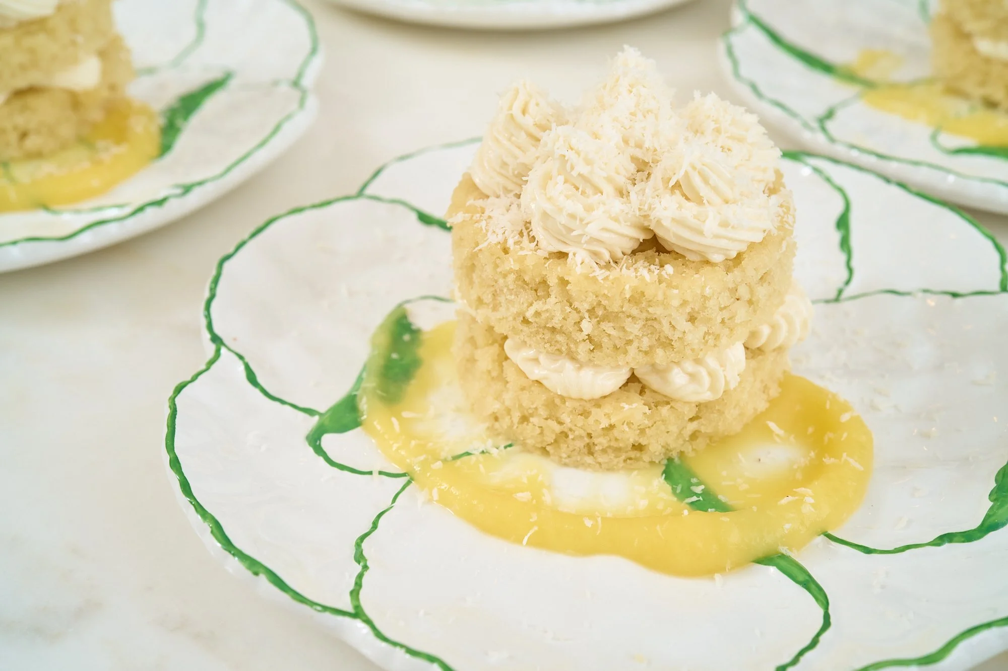 Close-up of a small yellow cake topped with white frosting and grated white chocolate, on a white plate with green accents and yellow sauce.