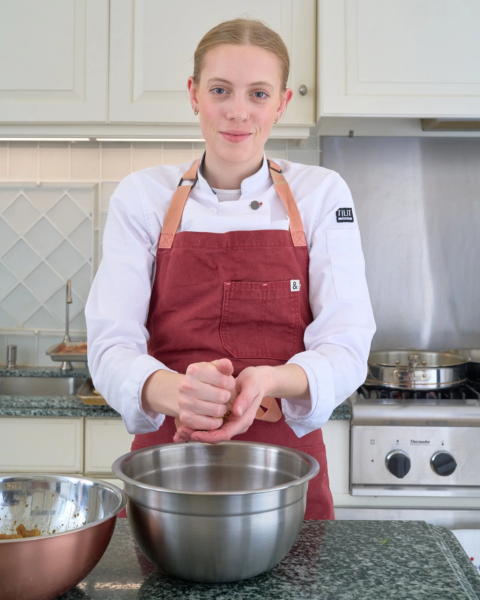 A young woman with blonde hair and blue eyes, wearing a red apron and white chef's jacket, is standing in a kitchen preparing food with her hands over a large stainless steel mixing bowl.