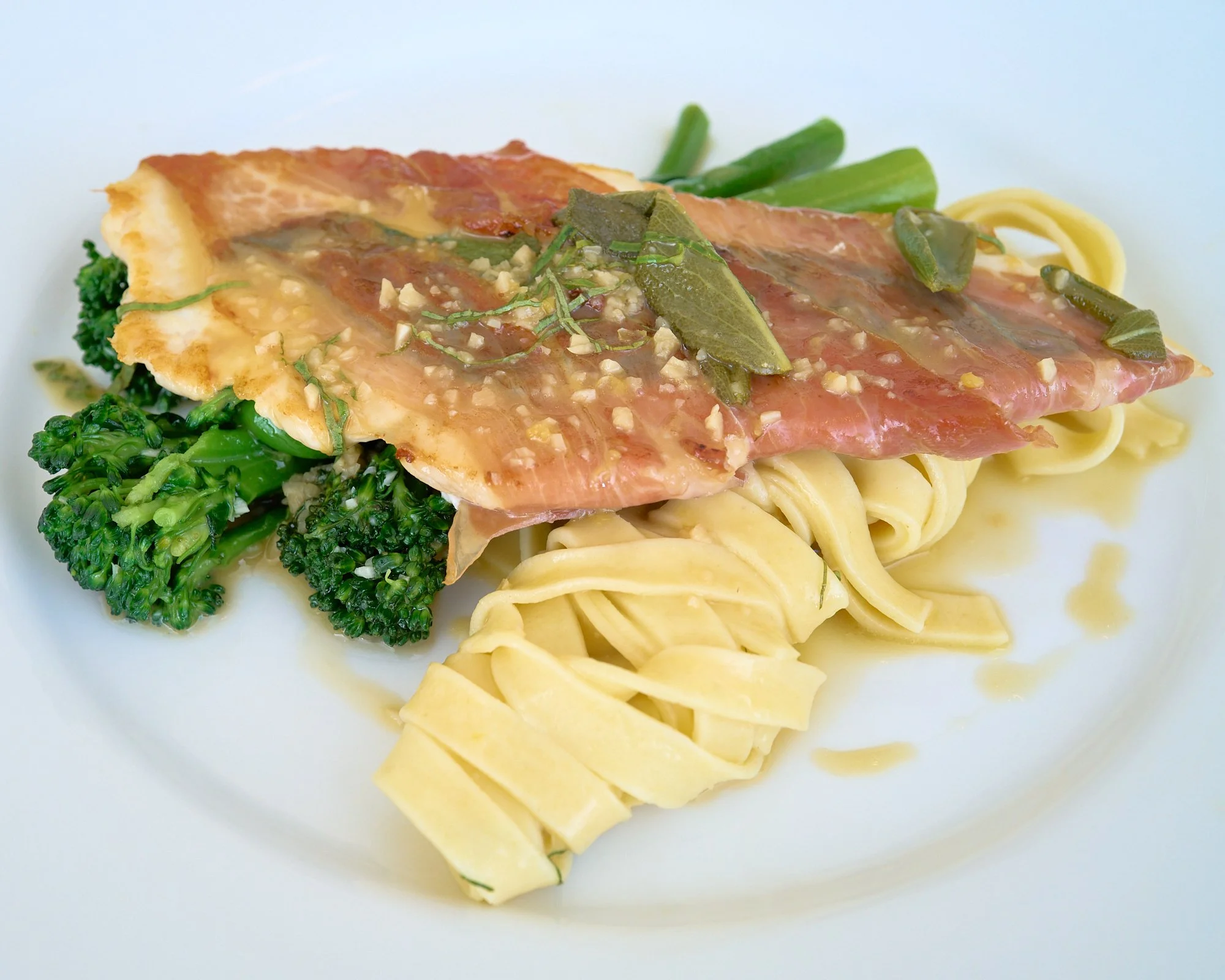 Plate of spaghetti with green vegetables, topped with cooked fish fillets garnished with chopped garlic, herbs, and sliced green peppers.