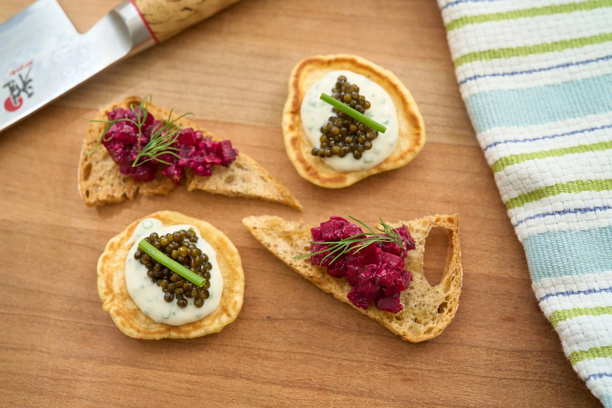 Close-up of assorted canapés on a wooden surface, topped with cream, caviar, herbs, and beet relish, with a striped cloth and a tube of caviar nearby.