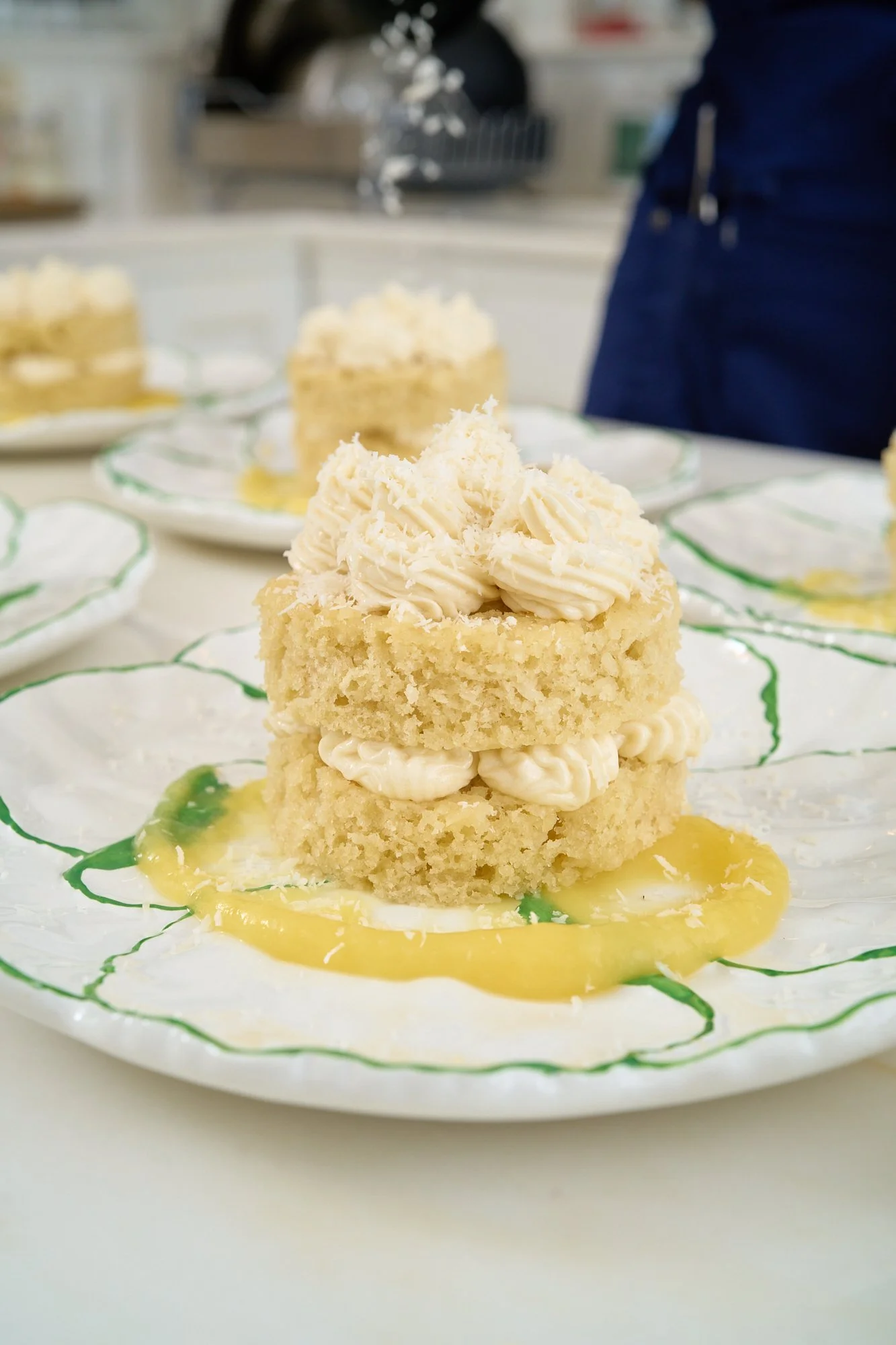 Layered vanilla cake with white frosting and grated coconut, on a decorative plate with green accents, with lemon sauce underneath.