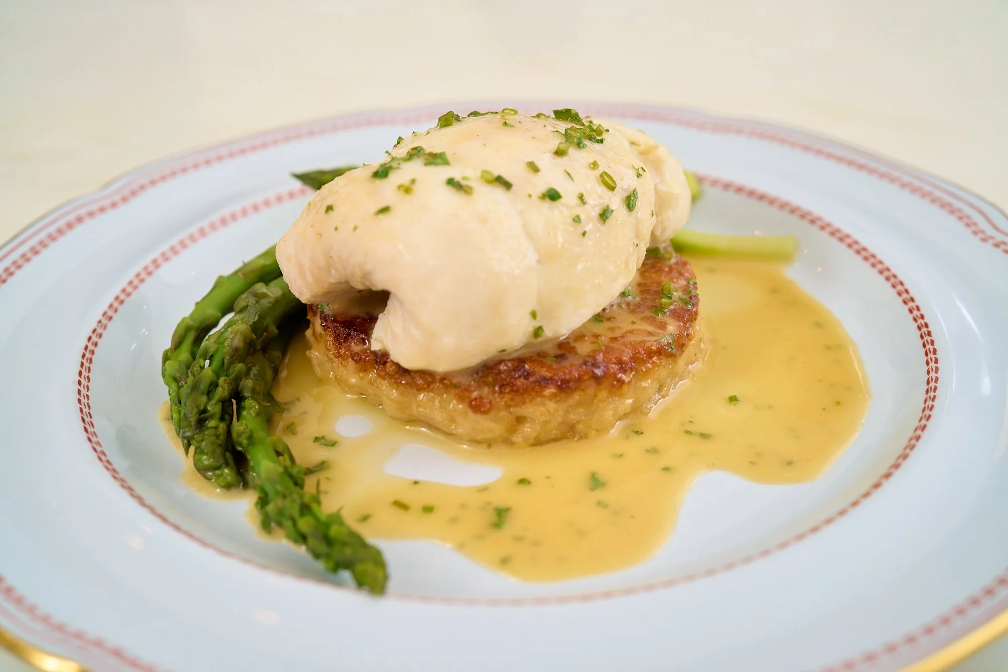 A plated gourmet dish featuring a seared crab cake topped with a dollop of creamy sauce and garnished with chopped herbs, accompanied by cooked green asparagus spears with a yellow sauce.