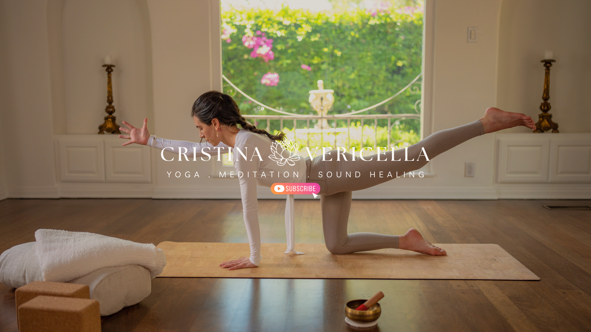 A woman practicing yoga indoors on a wooden floor with a view of a garden through a large window. She is on all fours with one leg extended behind her.