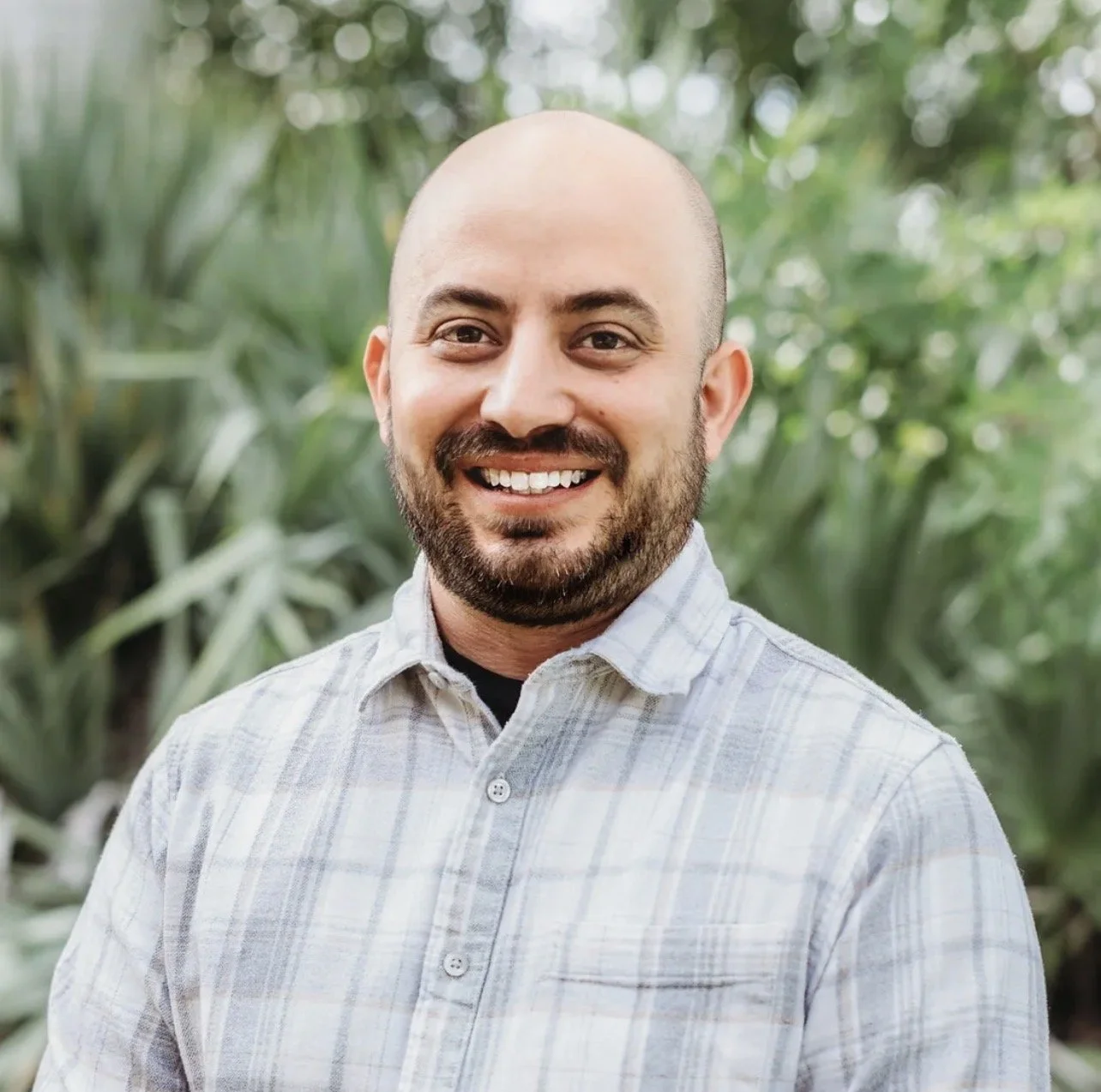 A smiling man with a beard, wearing a light-colored plaid shirt, standing outdoors with green foliage in the background.