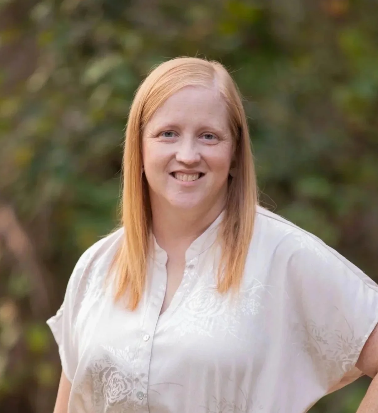 A woman with shoulder-length blonde hair smiling outdoors, wearing a cream-colored button-up shirt with a floral pattern, with a background of green foliage.