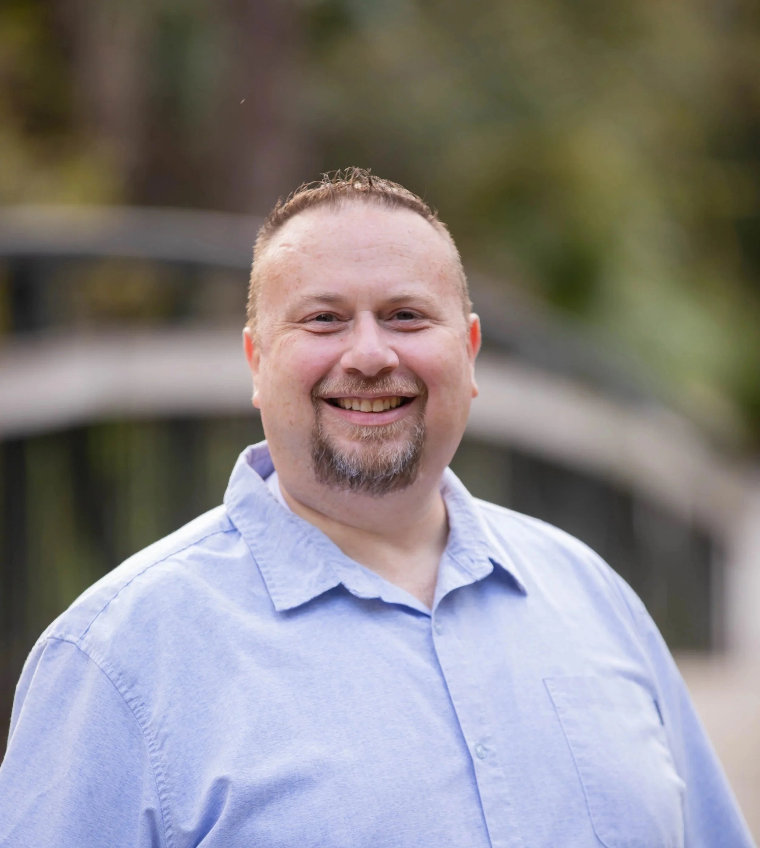 Close-up of a smiling man with short hair and a beard, wearing a light blue button-up shirt, outdoors with blurred greenery in the background.