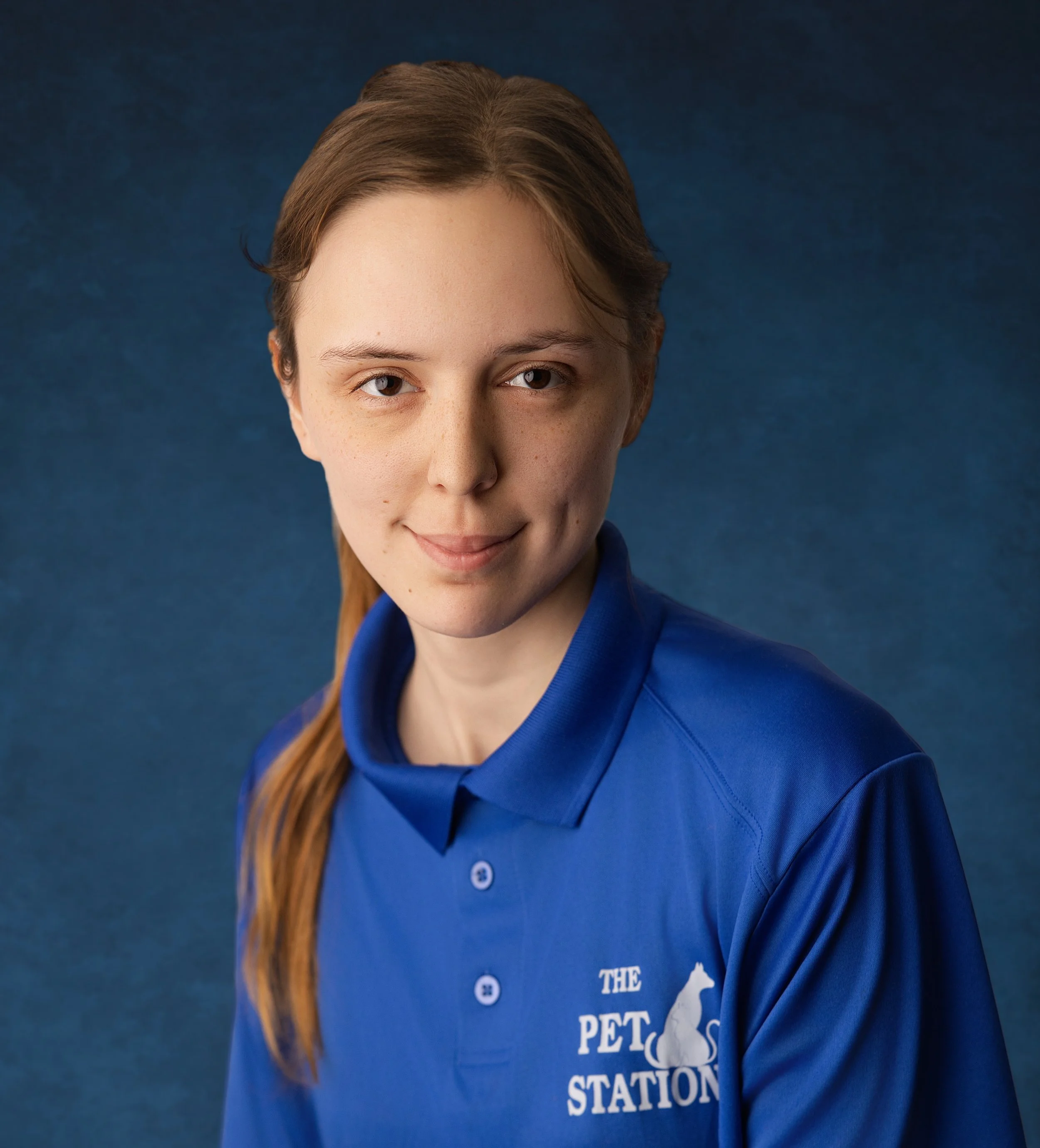 Abigail Culbertson - A young woman with red hair wearing a blue polo shirt with the logo and text for 'The Pet Station' on it, smiling slightly against a dark blue background.