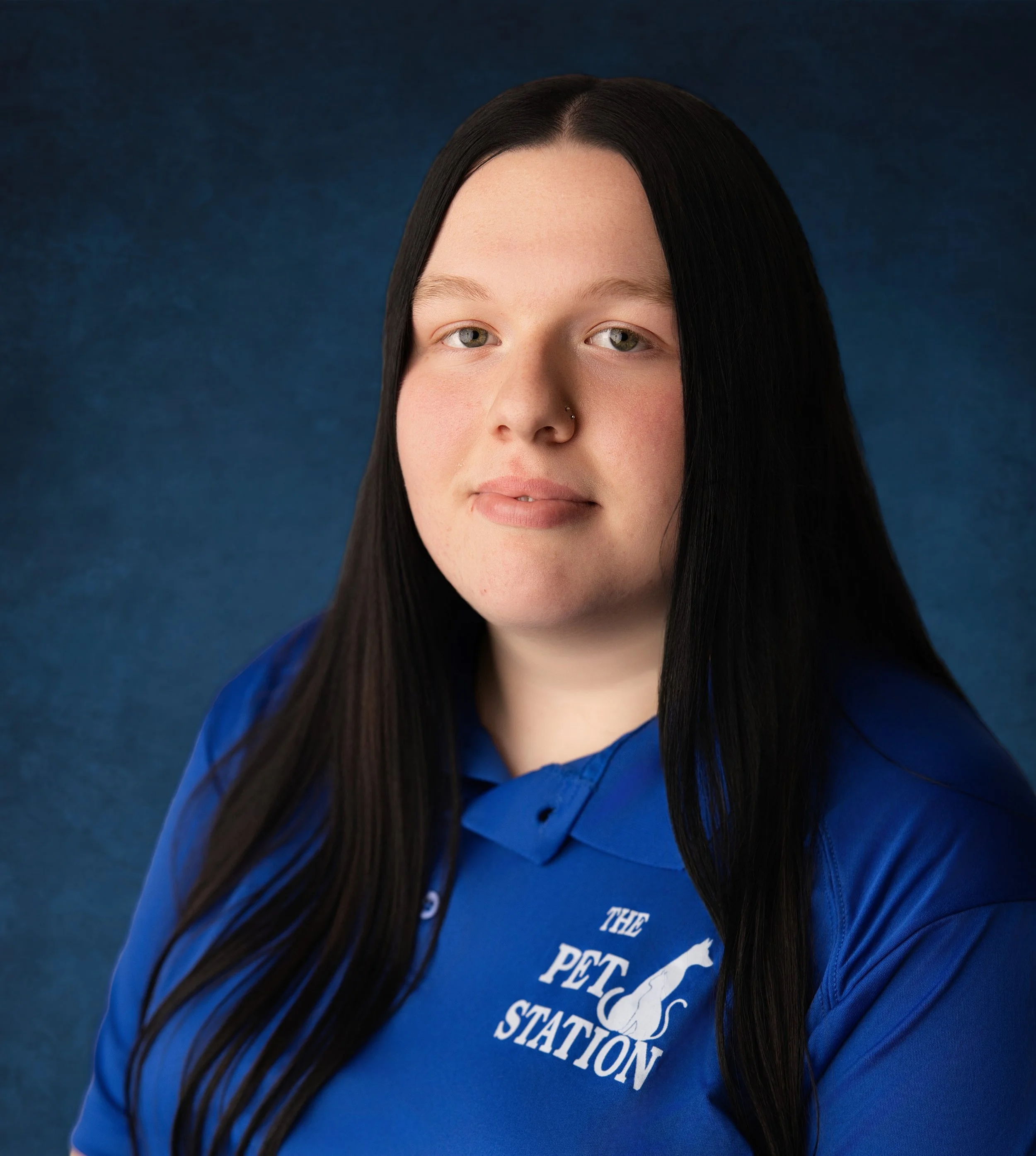 Kaylee Breaux - A woman with long dark hair, light skin, and a nose piercing, wearing a blue polo shirt with the logo 'The Pet Station' on it, posing against a dark blue textured background.