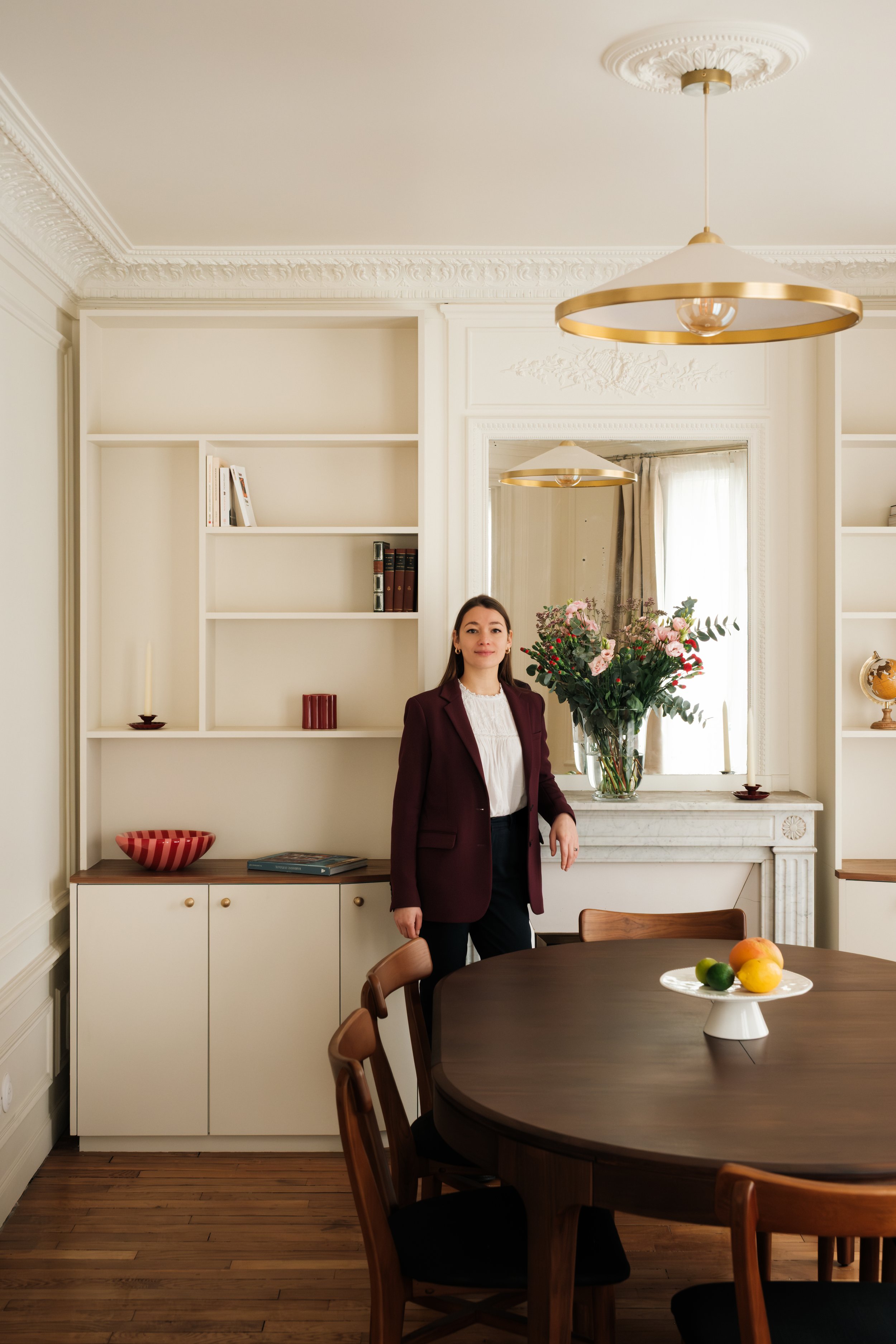 Klélia Delétang, architecte d'intérieur debout à côté d'une table en bois dans une salle à manger élégante avec des murs blancs, des étagères intégrées, une cheminée en marbre, un plafonnier design.