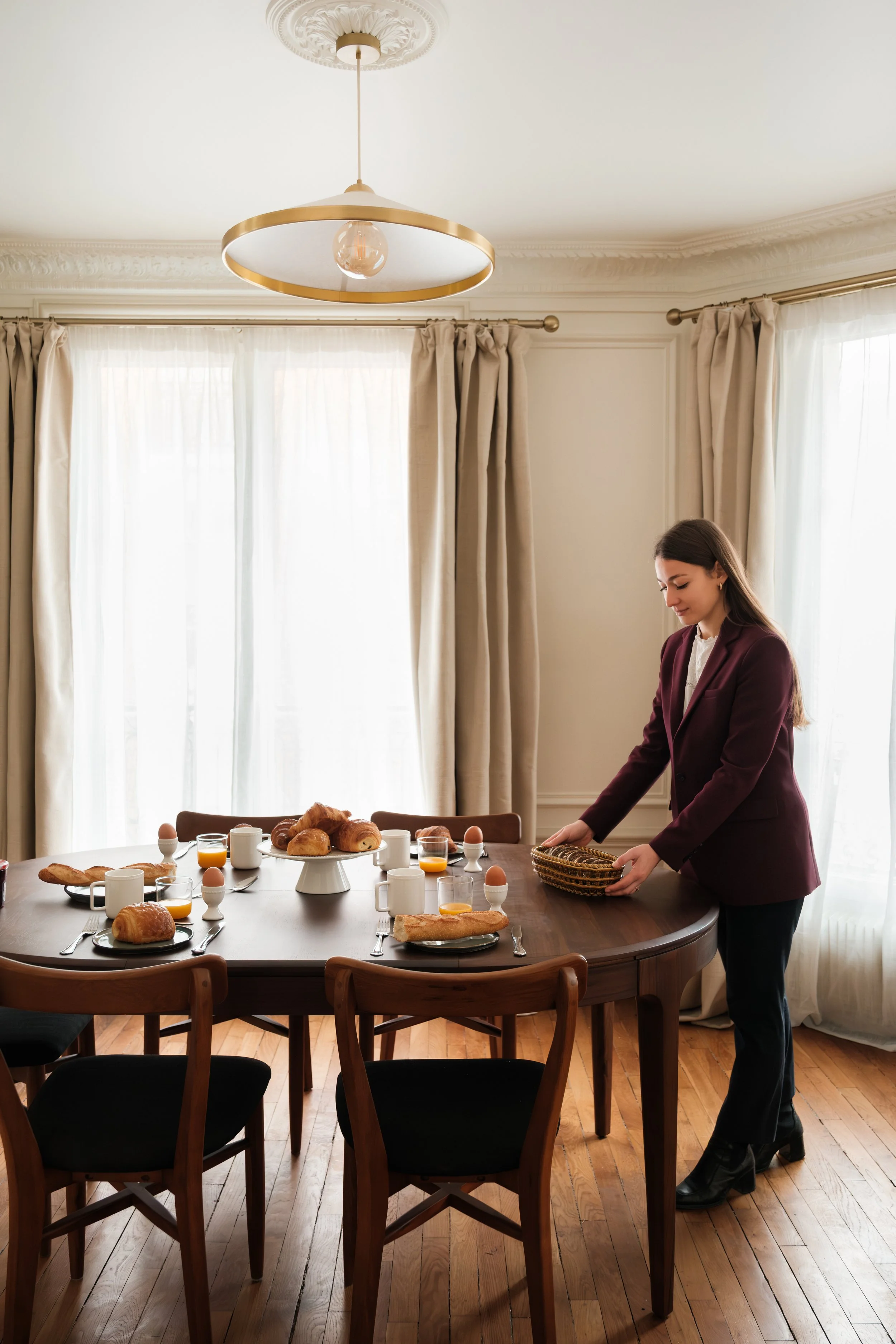 Une femme prépare une table pour le petit déjeuner dans une salle lumineuse avec des rideaux beiges, un chandelier moderne au plafond, et une table en bois carré avec des croissants, des œufs, du jus d'orange, et des tasses.