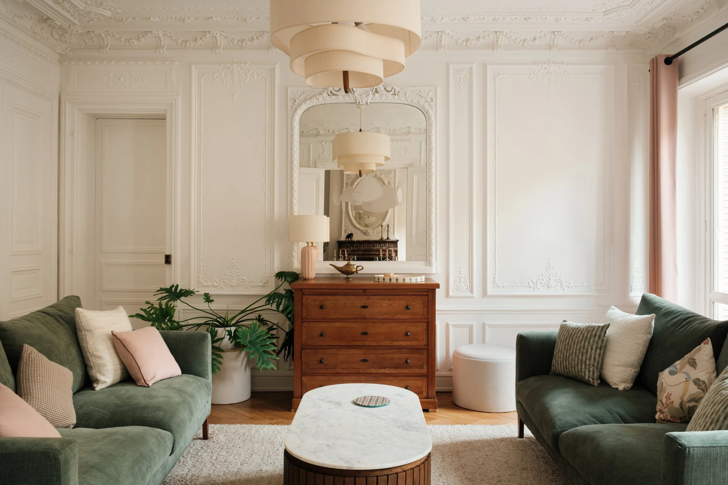 Salon avec deux canapés verts, un tapis beige, table basse en marbre, meuble en bois avec une lampe, un grand miroir et une fenêtre avec un rideau rose.