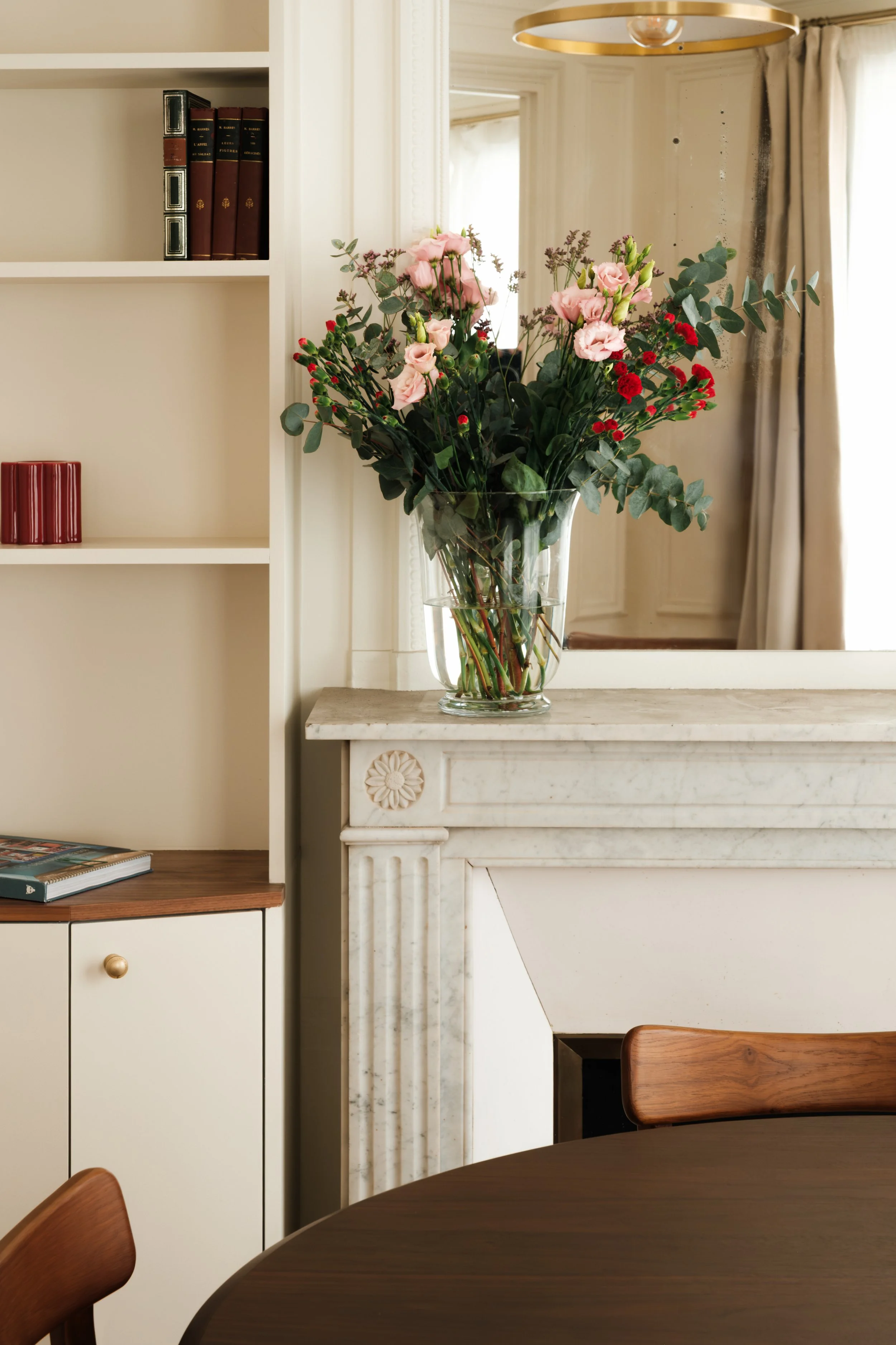 Un bouquet de fleurs dans un grand vase en verre placé sur une cheminée en marbre blanc dans une pièce lumineuse avec des rideaux blancs et des meubles en bois.