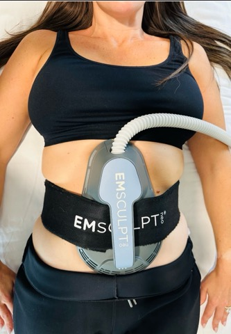 Woman undergoing EMSculpt body treatment with a device around her abdomen.