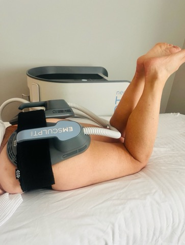 A person lying face down on a medical examination table with their legs elevated, connected to a medical device labeled "Emsculpt D" via a belt and hoses